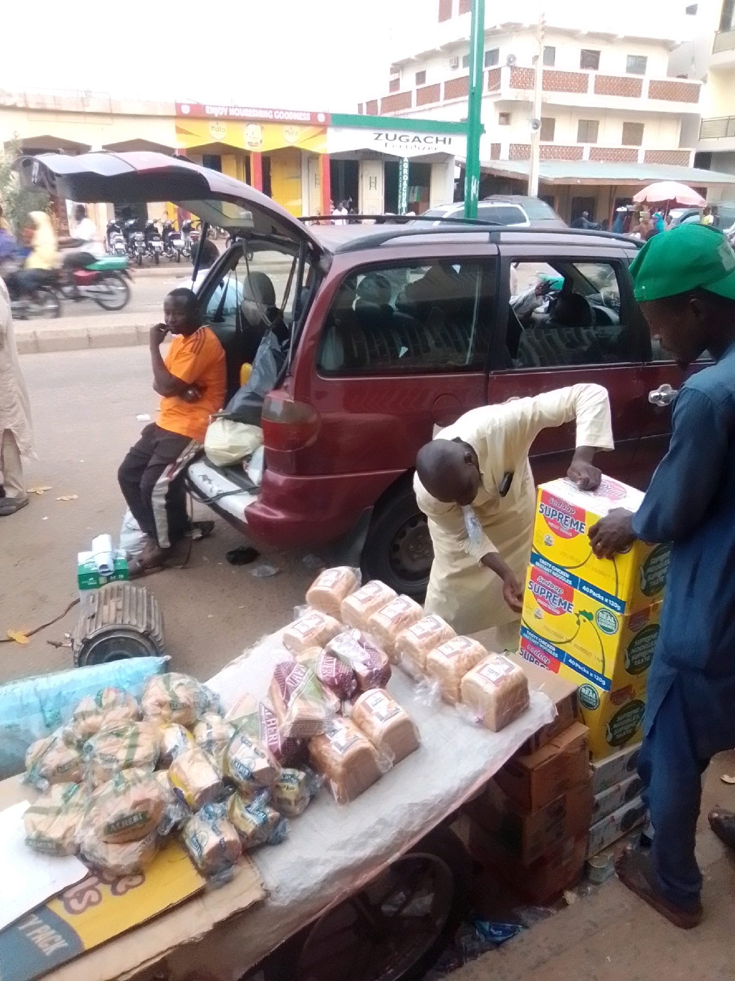 In Arawa, Nigeria, a street scene unfolds with a maroon van parked alongside a roadside stall. The van's rear hatch is open, revealing a partially visible interior and its contents. A man in an orange t-shirt and dark pants sits on the edge of the van's bumper, contemplating. Nearby, two men are engaged in moving a stack of large yellow boxes labeled "Supreme" and "Sadop Supreme" from the van to the stall. The stall is laden with loaves of bread, some individually wrapped and others in larger packaging, displaying labels with text such as "ALBERT" and "GOODNESS". Other items on the stall include a blue tarp and what appears to be an industrial motor. In the background, buildings and a green pole are visible, suggesting an urban environment. The ground is paved, and scattered debris indicates the commercial activity of the area.