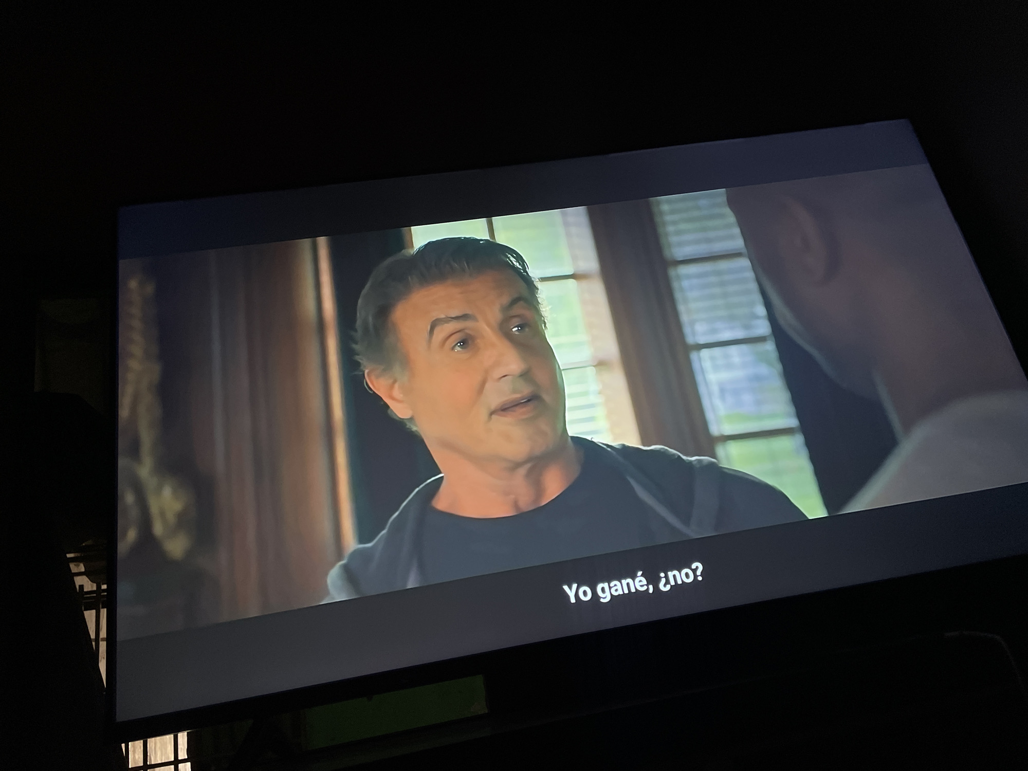 The image shows a screen displaying a scene with Sylvester Stallone. He is looking to the right and speaking. The text "Yo gané, ¿no?" is visible at the bottom of the screen. Stallone is wearing a black t-shirt and a gray jacket. The background includes window frames and what appears to be an interior of a house. Another person's ear is visible on the right side of the screen.