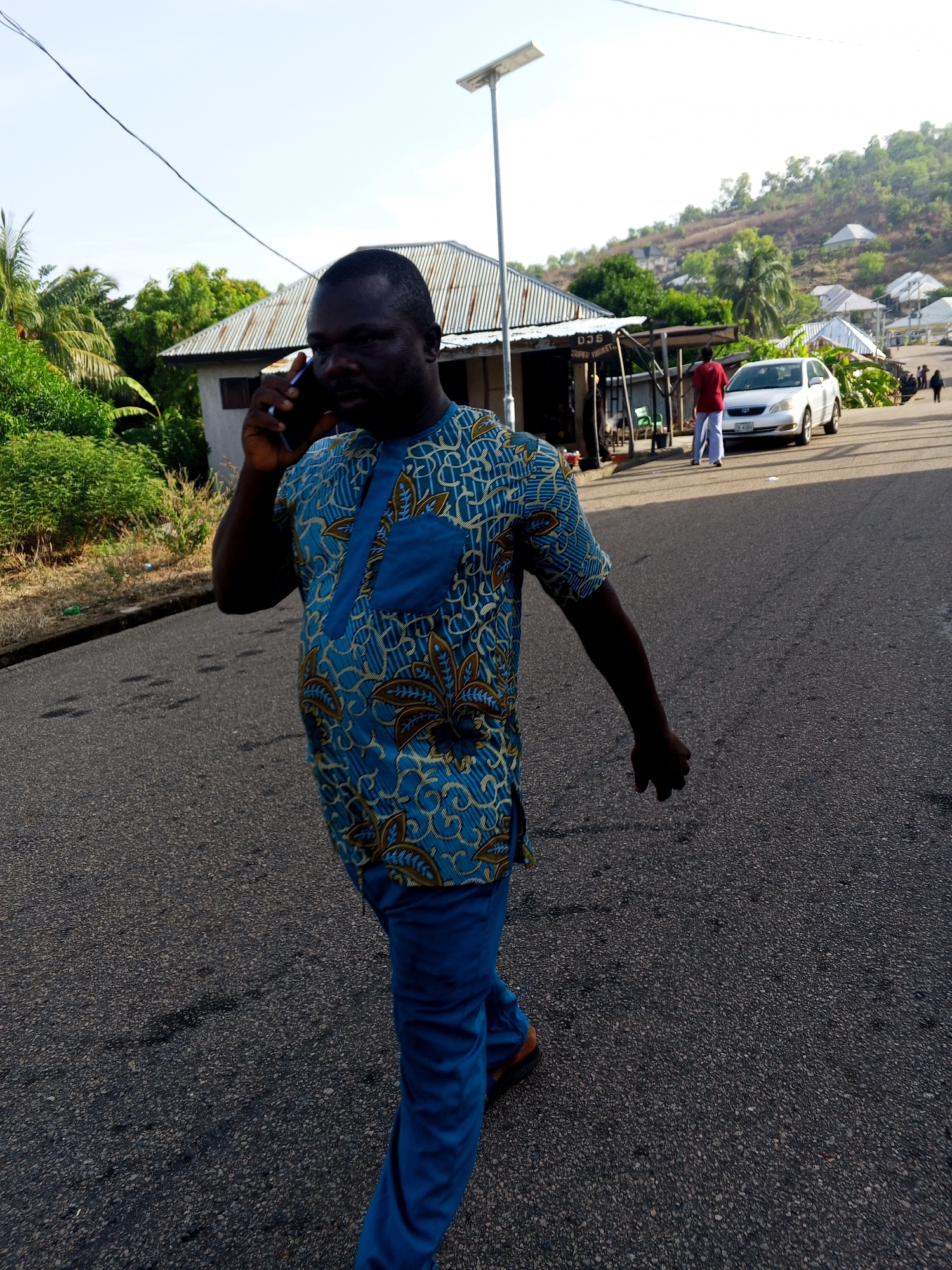 A man is walking on a paved road in Lokoja, Nigeria. He is wearing a blue patterned shirt and blue pants, and is holding a cell phone to his ear. In the background is a small building with a corrugated metal roof. A white car is parked on the side of the road, and a person in a red shirt and blue pants stands next to it. A utility pole stands next to the building. A small hill is in the far background with a few houses on it.