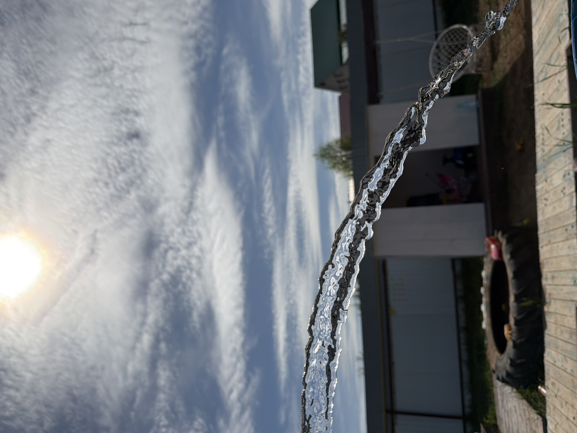 A low-angle shot presents a stream of water cascading against a bright, cloudy sky with the sun visible on the left. The water's surface is textured with ripples and highlights. The background includes a building and a tire leaning against a wooden structure. The exact location in Chaikovsky, Russia, cannot be determined from the image.