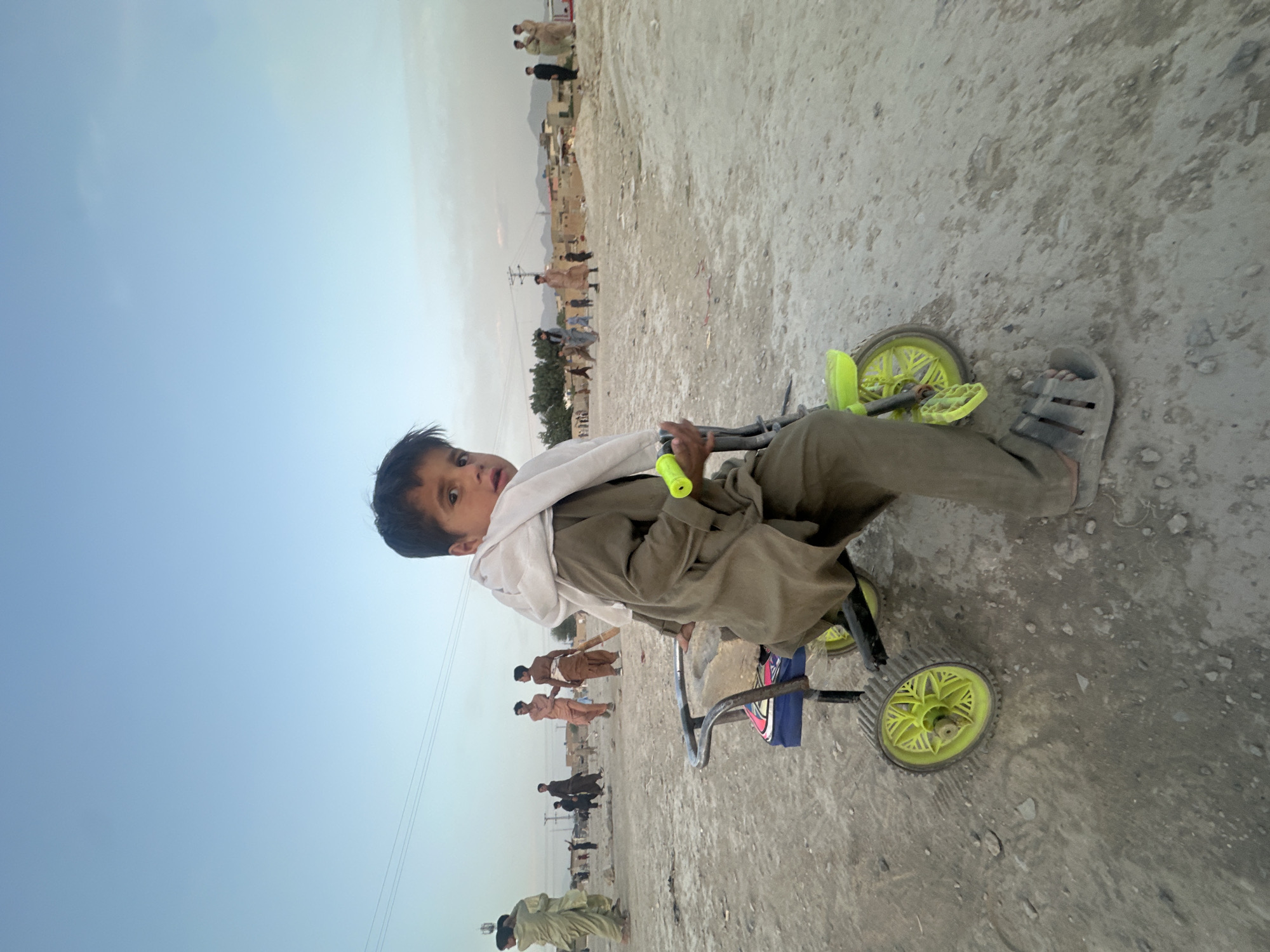 A child is on a tricycle on a dirt road in Quetta City Tehsil, Pakistan. The tricycle has lime green wheels. The child wears a brown garment and gray sandals and is draped with a white cloth. A group of people can be seen in the background. The sky is pale blue.