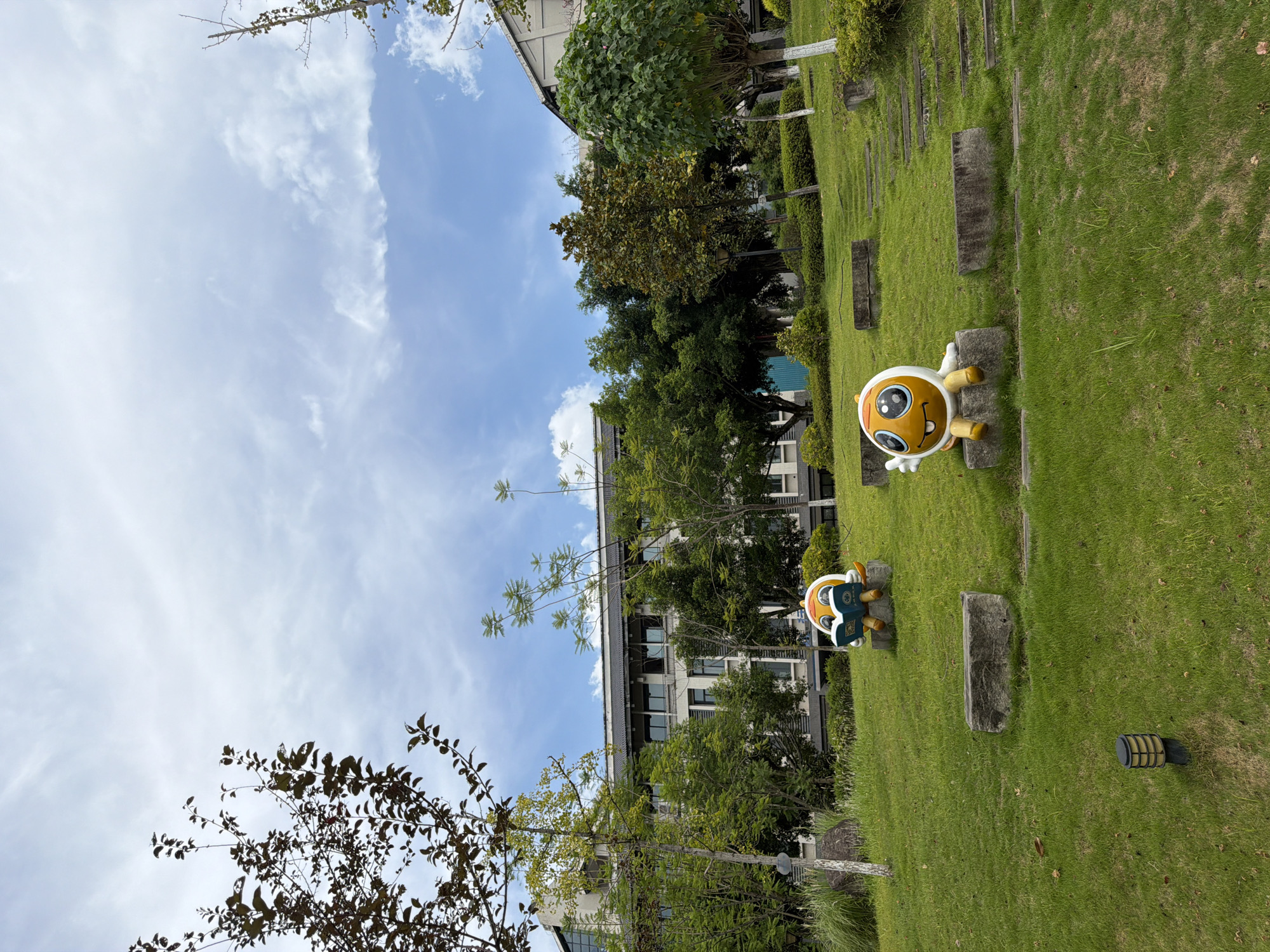 An outdoor, low-angle, medium shot captures a grassy slope with two cartoonish statues situated in the foreground. The statue on the left, a yellow, round figure with large, blue eyes and an orange head covering, is depicted reading a blue book with yellow pages. The statue on the right, also yellow and round with similar large blue eyes and an orange head covering, is sitting with its legs extended and one arm raised in a wave. Both figures are resting on square, concrete blocks. The environment is a well-maintained green lawn, with scattered fallen leaves and a single black lamp fixture near the bottom left corner. In the mid-ground, a pathway of concrete steps ascends the slope. Behind the statues and the slope, a modern building with multiple windows and a dark, tiled roof stands amidst lush green trees. The sky above is a mix of blue and white clouds, with the sun casting diffused light. The scene is likely set in an urban park or campus environment.