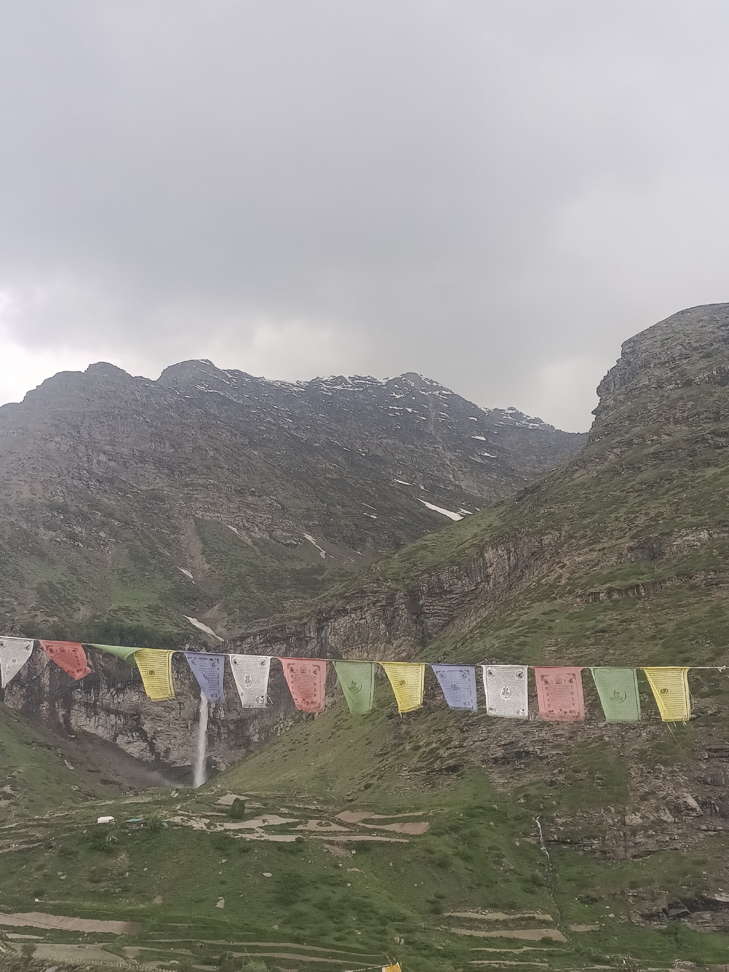 The image shows a scenic mountain landscape with prayer flags strung across the foreground. In the mid-ground, a waterfall cascades down a rocky cliff face, surrounded by lush green slopes. The mountains are imposing and rugged, with patches of snow visible near their peaks. The sky is overcast, contributing to a dramatic and somewhat somber atmosphere. The prayer flags are colorful, adding a touch of vibrancy to the natural setting. Small structures, possibly dwellings or rest stops, are visible on the lower slopes near the waterfall. The scene is located in Yamling, India, a region known for its natural beauty and spiritual significance, often visited by trekkers and pilgrims.