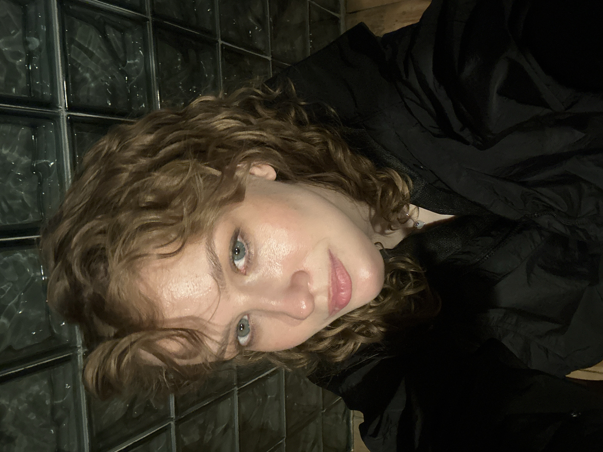 A close-up, high-angle selfie shows a fair-skinned woman with wavy, light brown hair and blue-green eyes. She is wearing a black jacket and looking towards the camera. Her face is illuminated, and there is a slight shine on her forehead and cheeks. The background consists of a dark, textured wall, possibly made of glass blocks. The lighting is dim, creating shadows and highlighting her features.