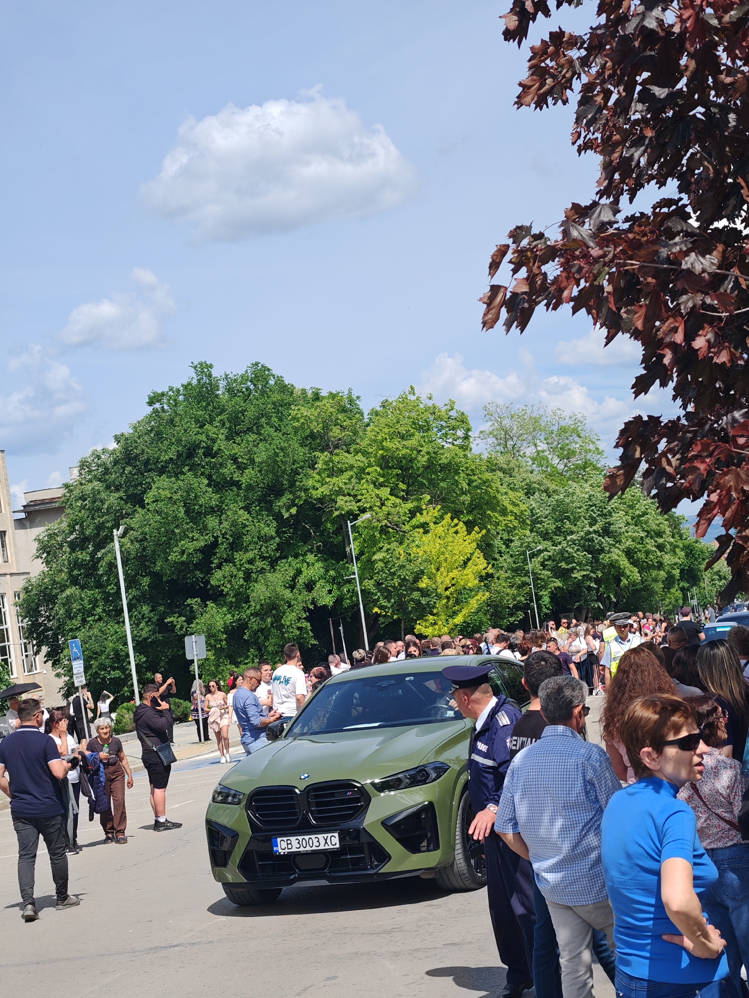 The image depicts a crowd of people gathered around an olive-green BMW X6 M car with a license plate that reads "CB 3003 XC". A uniformed police officer is standing next to the car. The crowd appears to be watching or interacting with the car or the police officer. The setting appears to be an urban street. A tree with dark red leaves is in the right corner of the image. A building is visible to the left, with multiple windows. The sky is partially cloudy. Based on the visual features and the location of the license plate, the scene likely takes place in the city of Shumen, Bulgaria.