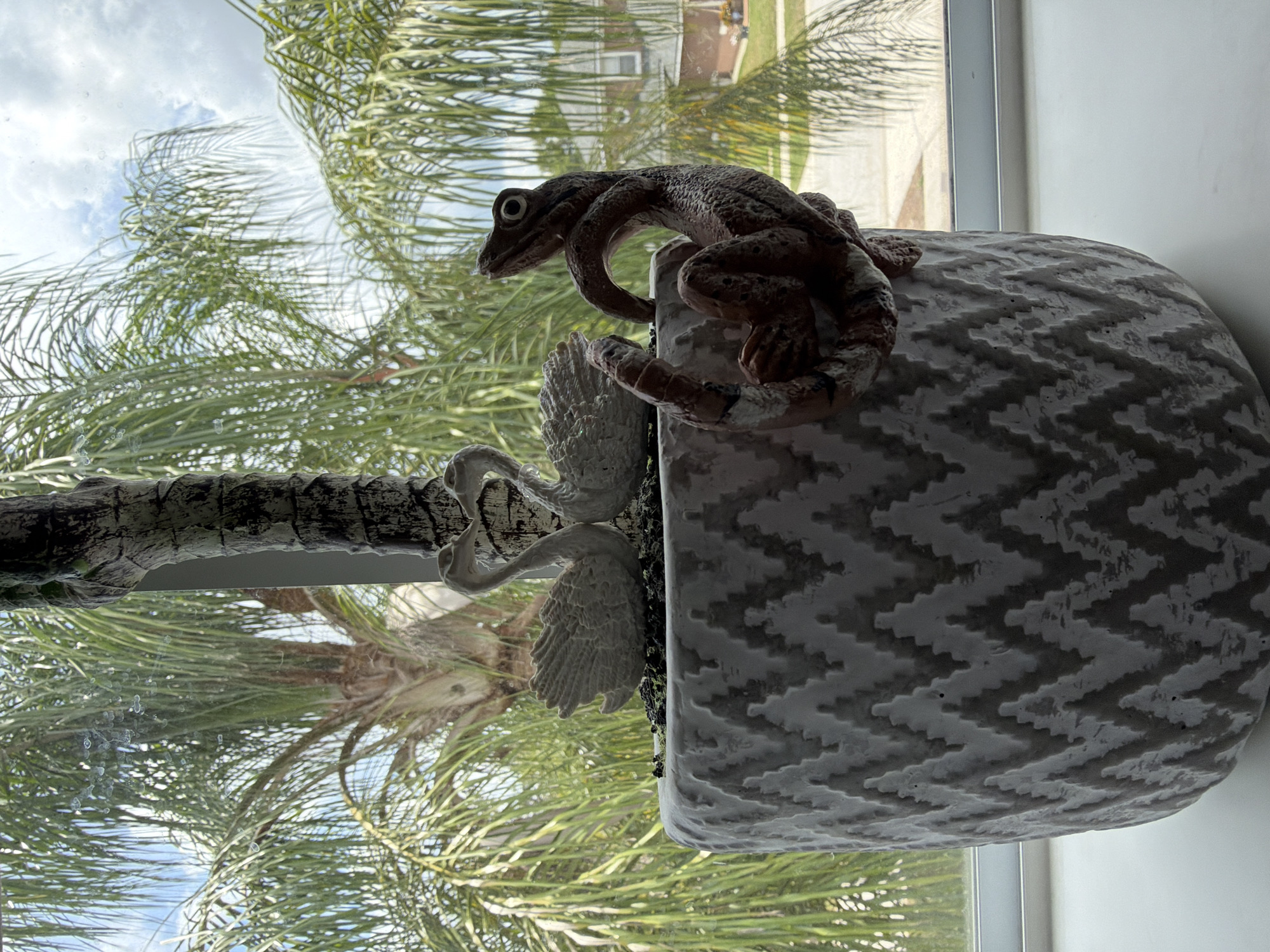 A close-up, eye-level shot captures a decorative artificial plant in a textured grey pot, positioned on a windowsill. The plant features a faux palm trunk, two white swan figures intertwined at the top, and a brown, lizard-like figurine clinging to the side of the pot. Through the window, a sunny sky with scattered clouds is visible, along with lush green palm trees and residential buildings in the distance, suggesting an outdoor setting in a warm climate. The scene is located in Riverview, United States of America.
