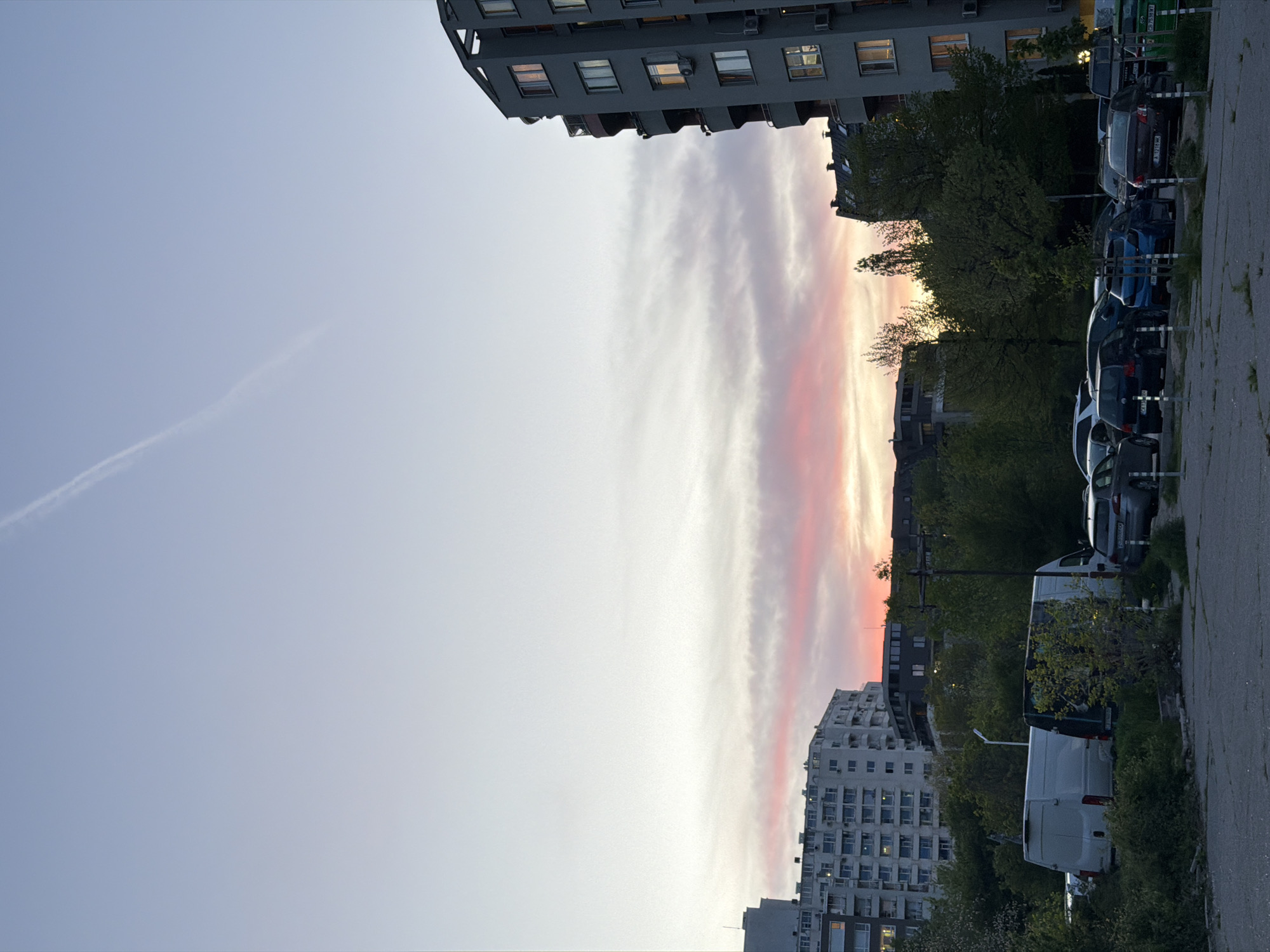 The image, rotated clockwise, captures an urban or suburban scene at either dusk or dawn, characterized by a colorful sky. The horizon displays a band of soft pink and orange hues, transitioning upwards into light grey and then a clear, pale blue. A single, thin white contrail stretches diagonally across the upper left portion of the blue sky.
The landscape is dominated by multiple multi-story apartment buildings. On the upper left, a dark-colored building with a distinct rounded corner and several illuminated windows is prominent. To the lower right, a large, older, light-colored apartment block with numerous balconies and windows is visible. Other buildings with various architectural styles are scattered throughout the mid-ground.
In the foreground and mid-ground, numerous vehicles are parked along what appears to be a street or parking area. These include various sedans, SUVs, and vans, mostly dark in color, though a white van is clearly visible in the lower right. Several trees and bushes are interspersed between the buildings and parked cars, providing greenery to the scene. The paved ground in the foreground on the right appears to be a sidewalk or a part of the parking lot.
Visible text includes license plates on two cars: "CA 2144 MH" on a dark blue car and "CA 3034 MA" on a gray car. The "CA" prefix on these plates confirms the location as Sofia, Bulgaria, as provided in the context. The overall impression is one of a quiet urban residential area during the gentle light of sunset or sunrise.