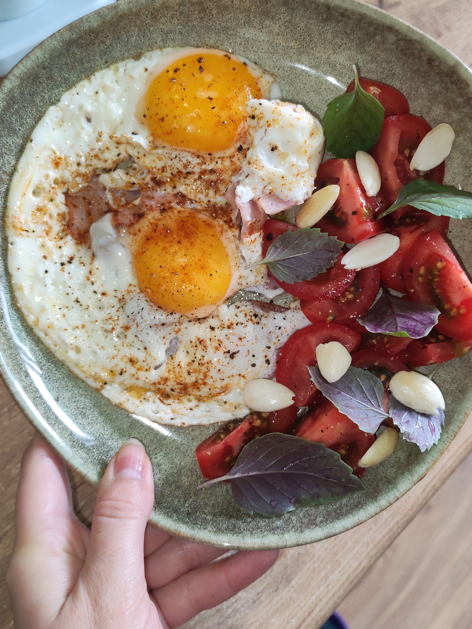 A meal is presented on a round, textured, earthy-green ceramic plate, situated in the city of Izhevsk, Russia. The left side of the plate features two fried eggs with distinct bright yellow-orange yolks and fully cooked whites, seasoned with dark and reddish-brown spices. Portions of cooked meat, possibly bacon or ham, are visible beneath and between the eggs. The right side of the plate holds a salad composed of roughly chopped red tomatoes, adorned with several fresh basil leaves, varying in green and purplish hues, some exhibiting small water droplets. Multiple blanched, whole almonds are scattered across the tomato salad. A fair-skinned human hand, with a short thumbnail visible on the thumb, is positioned in the lower left, holding the rim of the plate. The plate rests on a light brown wooden surface with a visible grain pattern. A fragment of a white object is partially discernible in the upper left background.