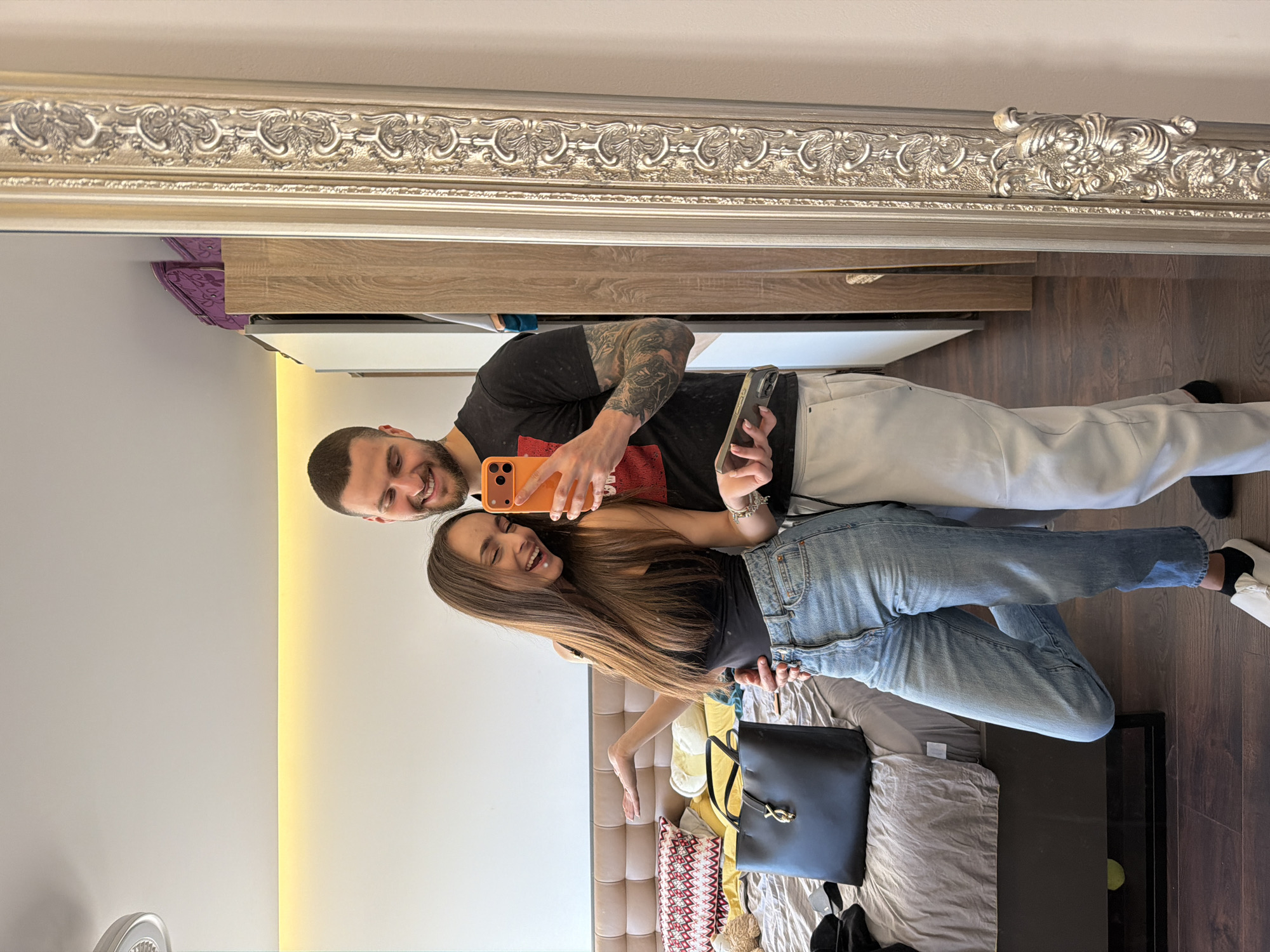 A man and a woman are taking a selfie in front of a mirror. The man is wearing a black t-shirt, white pants, and has tattoos on his left arm. He is holding an orange smartphone. The woman is wearing a black top and blue jeans. They are both smiling and appear happy. They are in a room with a bed, and the mirror has an ornate silver frame. A black handbag and other personal items are on the bed. The location is most likely an apartment or hotel room. The city of Sofia, Bulgaria is not evident from the image.