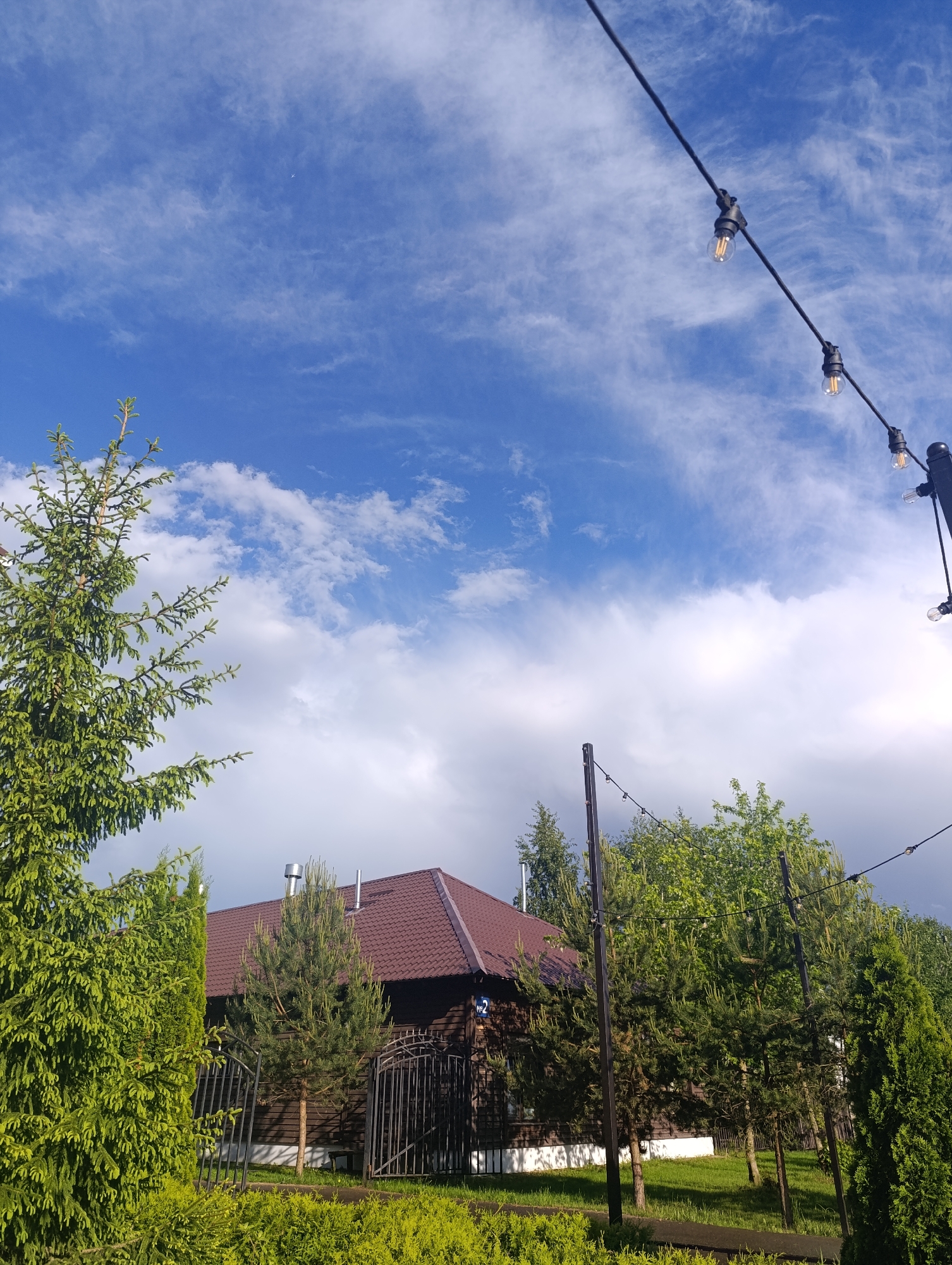 The image shows a house partially obscured by trees, under a sky of blue and white clouds. The house has a brown roof and dark wood siding, with metal chimney stacks. A black metal fence is visible in front of the house. String lights hang diagonally across the upper right portion of the frame. The location is Unknown, Unknown.