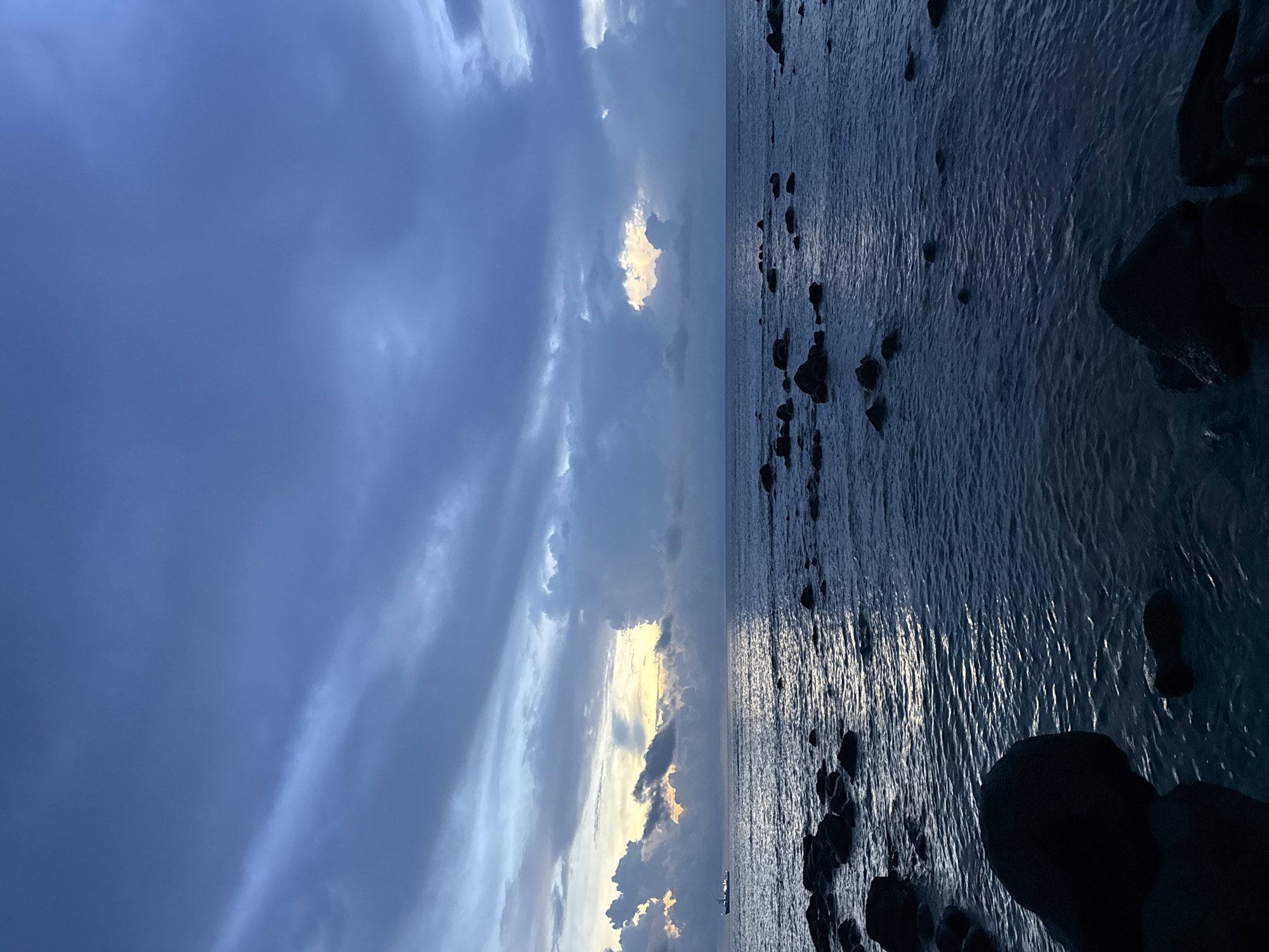 The image depicts a twilight seascape with a focus on the ocean, rocks, and sky. The foreground is dominated by dark, irregularly shaped rocks scattered along the waterline, some partially submerged in the gentle waves. The water itself is a deep, muted blue, reflecting the subdued light of the setting or rising sun. In the distance, a faint horizontal line suggests the horizon where the sea meets the sky.
The sky is a prominent feature, filled with dramatic clouds. The lower portion of the sky shows patches of warm, golden light breaking through darker, more ominous clouds, suggesting a dramatic transition in weather or time of day. A small, distant vessel, possibly a ship or a large boat, is visible on the left side of the frame, moving across the water. The overall mood is serene yet tinged with the dramatic atmosphere of twilight. The location is the coast of Lý Sơn Special Administrative Region, Vietnam, known for its volcanic rock formations and coastal scenery.