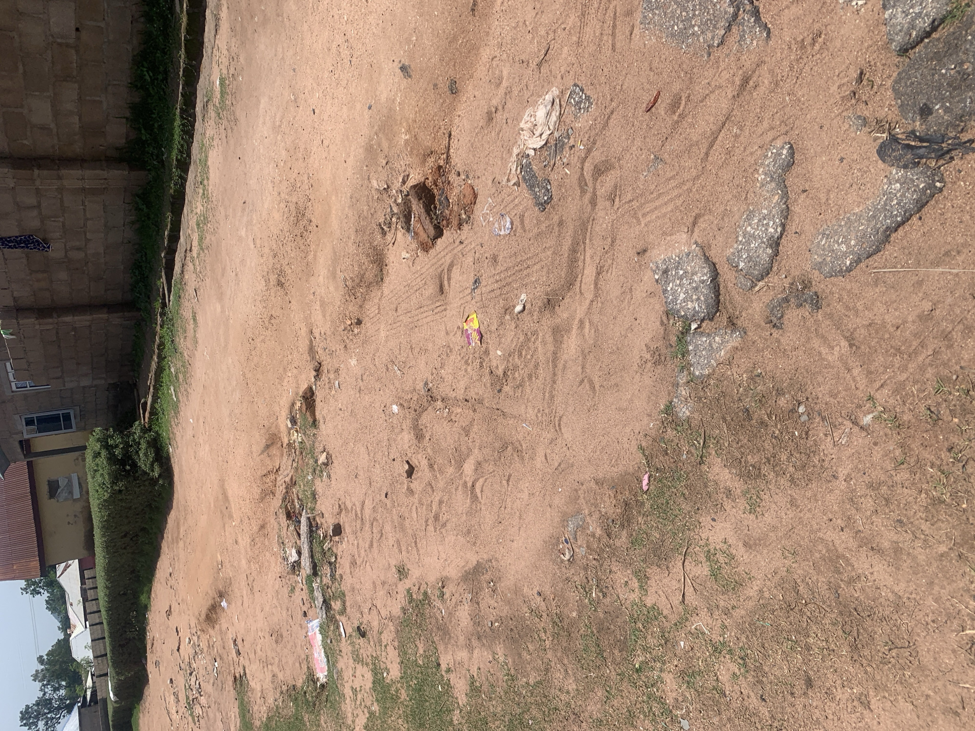 The image depicts a dirt road with scattered debris in Jauro Yinu, Nigeria. The road surface is uneven, composed of reddish-brown soil and sand, with visible patches of broken asphalt and small stones. There is evidence of tire tracks imprinted in the sand. Various pieces of litter are strewn across the ground, including plastic wrappers, a torn piece of white cloth, and broken bricks. In the background, a low, dense green hedge runs along the side of the road, separating it from buildings. These buildings appear to be constructed with concrete blocks and some have corrugated metal roofs. The sky is visible in the upper portion of the image and appears to be overcast or hazy. No people are visible in the scene.