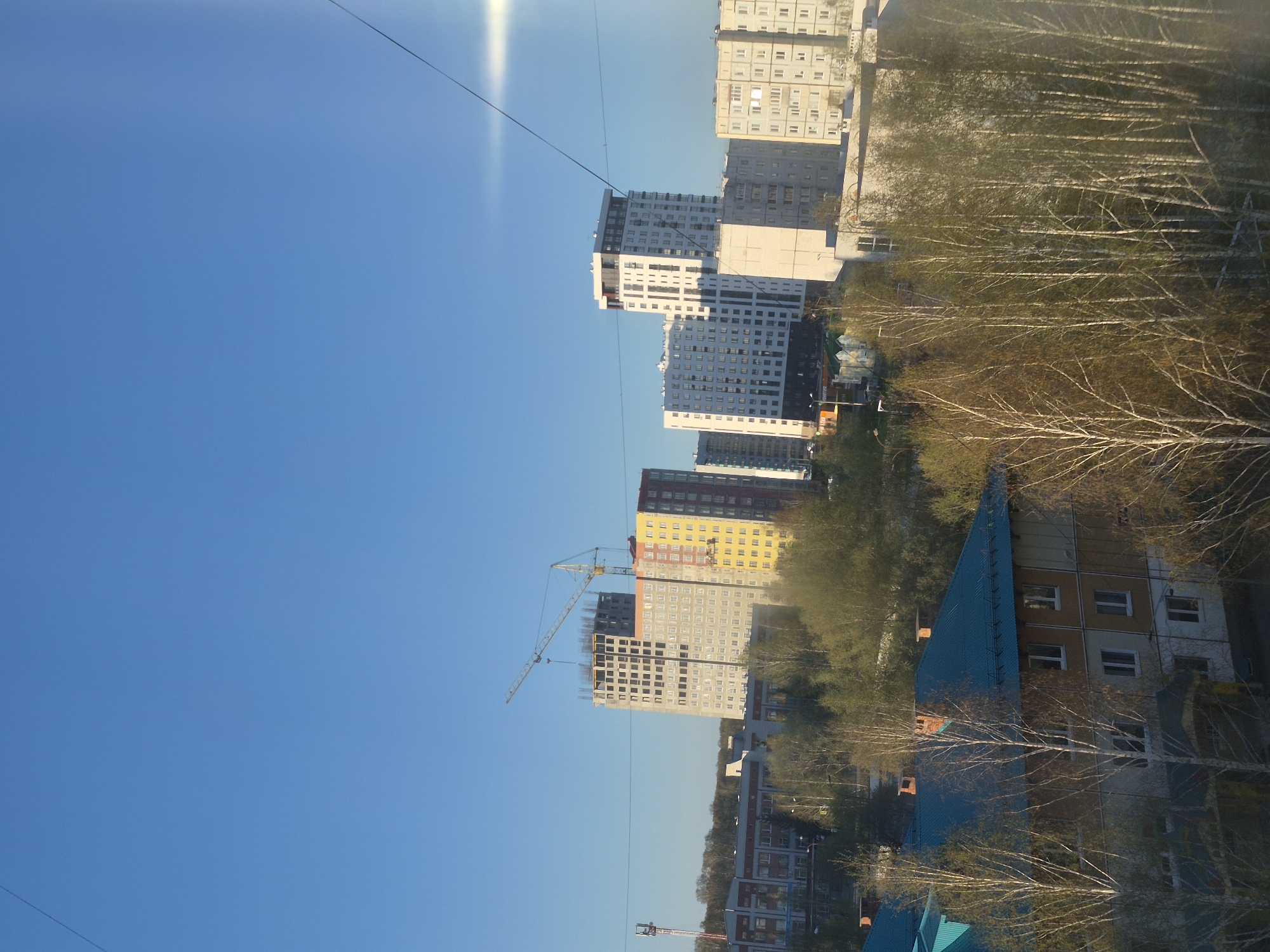 The image presents an urban landscape, likely in Tobolsk, Russia, based on the architecture and vegetation. It showcases a combination of older and newer buildings, including high-rise apartment complexes under construction. A construction crane is visible near one of the buildings. The foreground features trees, possibly birch, with bare branches suggesting late autumn or early spring. A clear, blue sky dominates the upper portion of the image. Power lines are visible stretching across the sky.