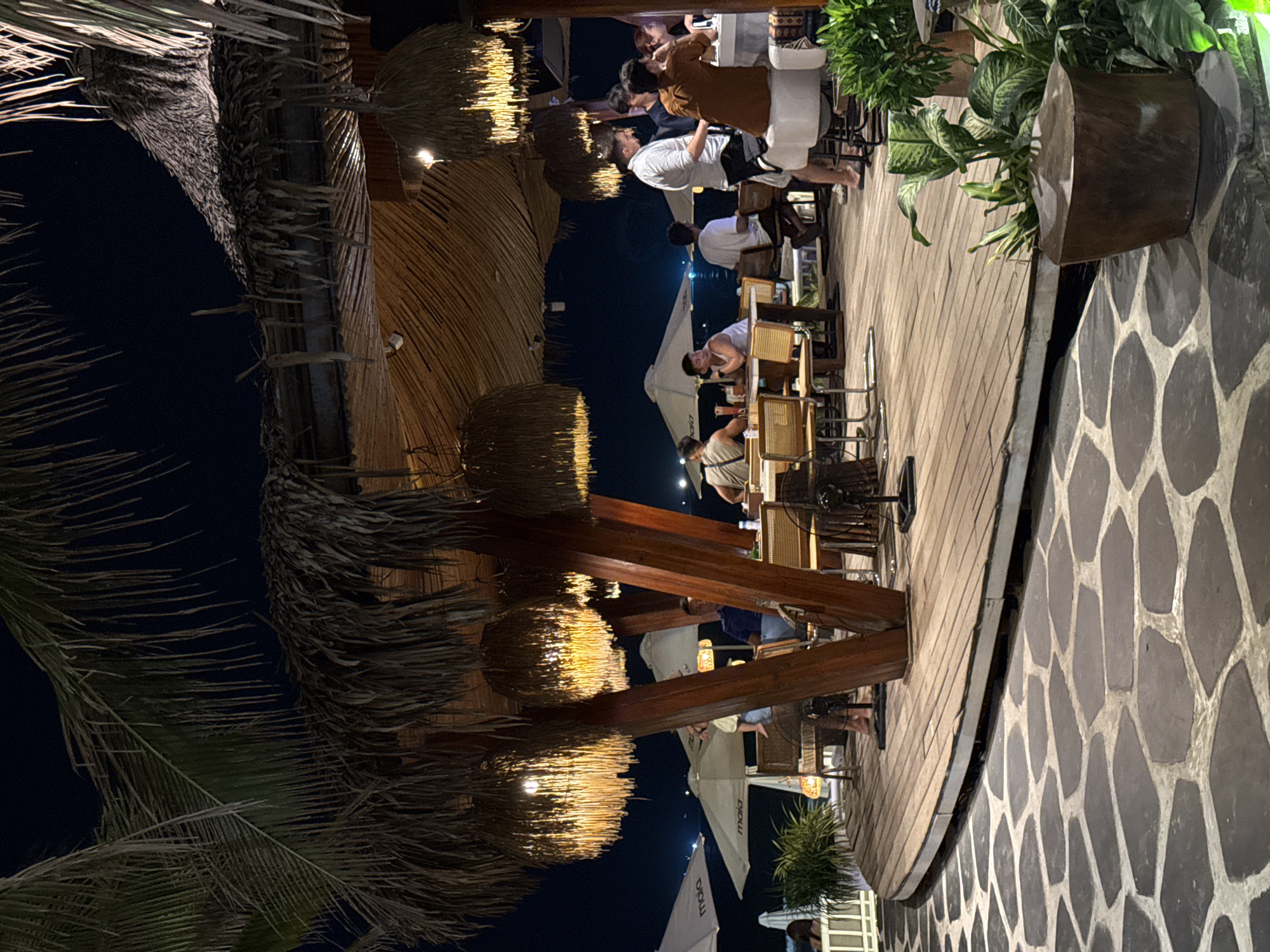 This image captures an evening scene at an outdoor restaurant or bar in Da Nang, Vietnam. The setting features a rustic, tropical design with a large thatched roof supported by robust wooden beams. Warm light emanates from multiple spherical, woven lanterns hanging from the roof structure, illuminating the area.
Several individuals are present, seated at various tables on a raised wooden deck. They appear to be patrons, casually dressed, engaging in conversation, dining, and drinking. Some are seen interacting, while others are looking towards their tables. The ambiance is relaxed and social.
The floor surrounding the wooden deck is paved with large, irregularly shaped stone tiles. Tropical vegetation is prominent, with palm fronds visible in the darker foreground and background, and potted plants adding greenery to the deck area. A white umbrella, partially visible towards the center-right, has the text "MAIA" printed on it. The dark sky confirms it is nighttime, and the artificial lighting provides the primary illumination for the scene, enhancing the inviting atmosphere of the establishment.