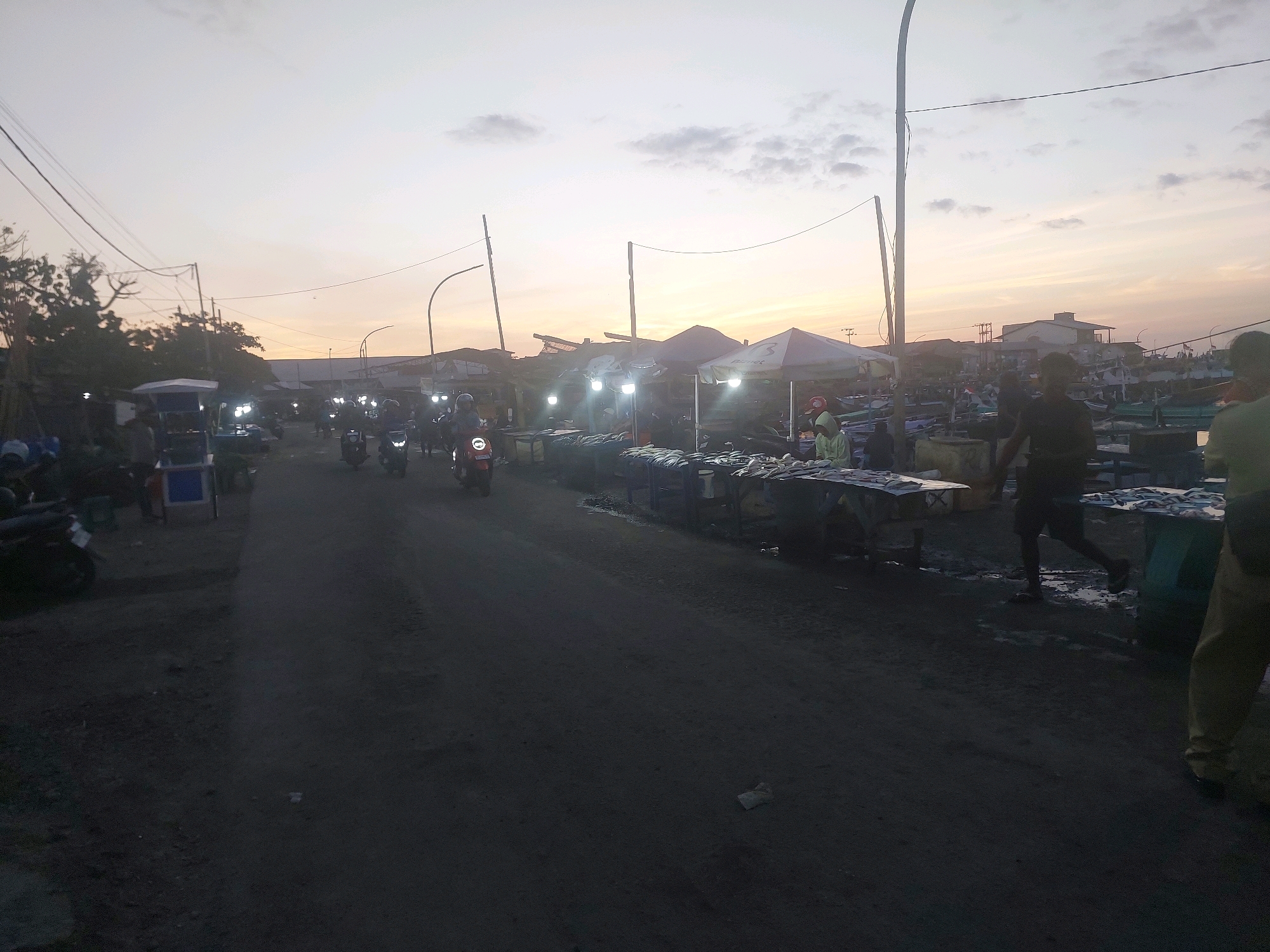 The image depicts a bustling street market at dusk, possibly near a fishing harbor, in Kupang, Indonesia. The sky is a soft blend of orange and gray, indicating either sunrise or sunset, with scattered clouds. In the foreground, a dark, unpaved road stretches into the distance, with several motorcycles parked or in motion on the left side.
The right side of the road is lined with market stalls covered by tents. These stalls display what appear to be fresh fish laid out on tables, suggesting a fish market. Several individuals are visible in and around the stalls, some appearing to be vendors or customers. One man, dressed in dark clothing, is running across the road. Another person, wearing a yellow shirt and brown pants, is walking away from the camera.
Further into the background, more market stalls and structures are visible, along with what could be fishing boats, hinting at a proximity to the sea. Streetlights are illuminated, casting a glow on the scene and highlighting the activity despite the fading light. The overall atmosphere is one of active commerce and community engagement in the twilight hours.