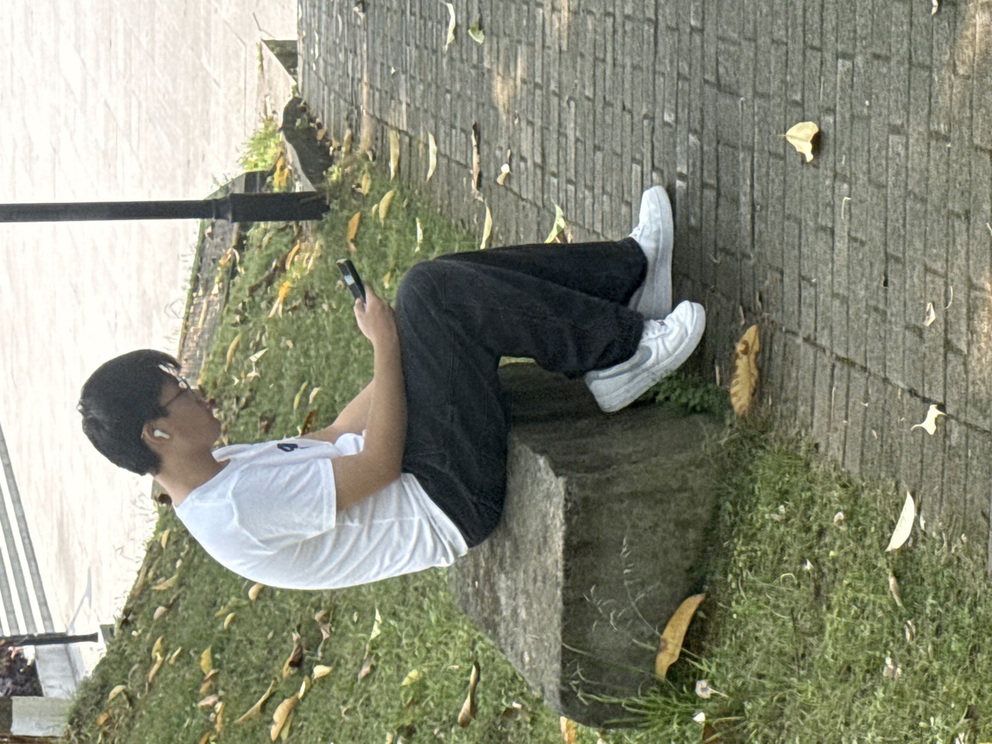 A person wearing a white t-shirt, dark pants, and white shoes sits on a stone ledge, holding a smartphone. They have short dark hair and wear glasses. A white earbud is visible in their left ear. The ledge is positioned beside a pathway made of grey rectangular stones. Grass and scattered yellow leaves are on the ground adjacent to the path. A black lamppost stands to the left of the person. The scene appears to be outdoors in a park or garden. While the location is specified as Jiangjin, China, there are no visual elements to verify the specific location.