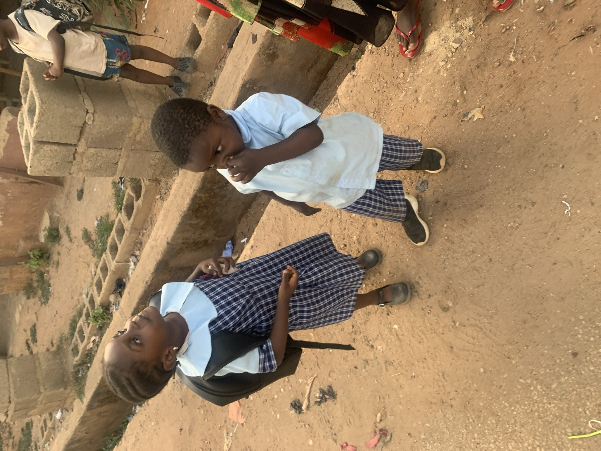 In New Stadium, Nigeria, two children in school uniforms stand on a dirt surface near a concrete structure. One child, dressed in a light blue top and plaid pants, touches their face. The other child wears a plaid dress with a light blue collar and carries a black backpack. Other people's legs are visible in the background. Debris is scattered on the ground.