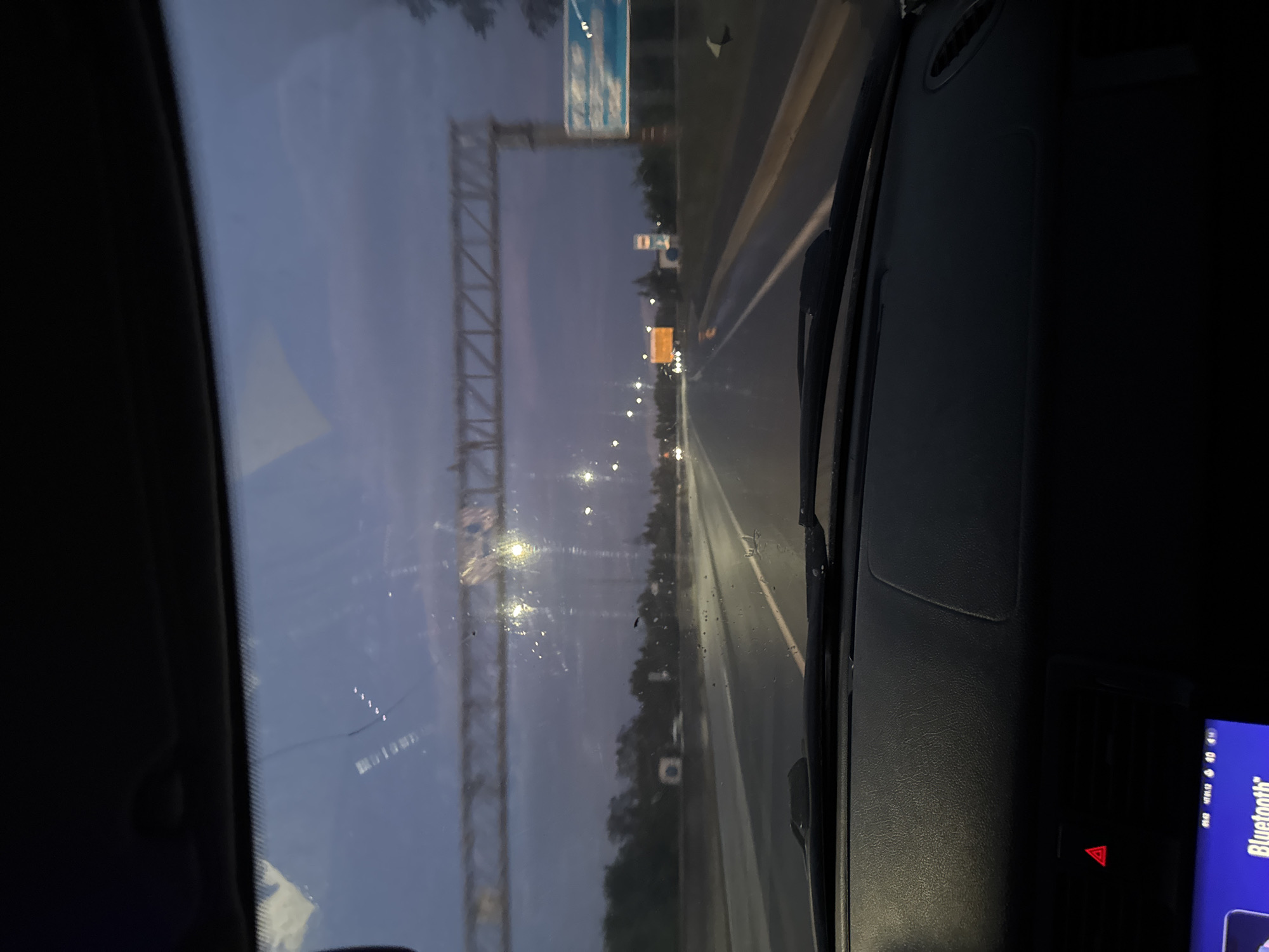 The image, taken from inside a vehicle, shows a view through the windshield of a highway at dusk. The road appears wet, suggesting recent rain. Overhead, a large metal structure, likely a gantry for traffic signs or lighting, spans the highway. In the distance, streetlights illuminate the road and the surrounding trees. A blue and white sign is visible on the right side of the highway. The dashboard of the car is visible in the foreground. The sky is a deep twilight blue with some clouds. There are no people or other vehicles clearly visible and identifiable in the image. The location is identified as the city of Perm, Russia.