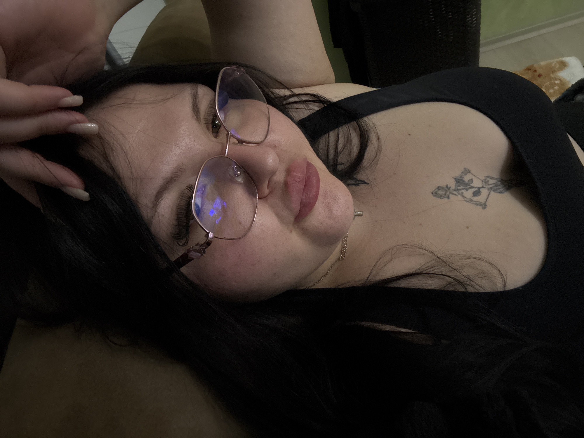 A woman with long dark hair is lying down, possibly on a sofa or bed. She is wearing glasses and a black top. A tattoo is visible on her chest. The location is unknown.