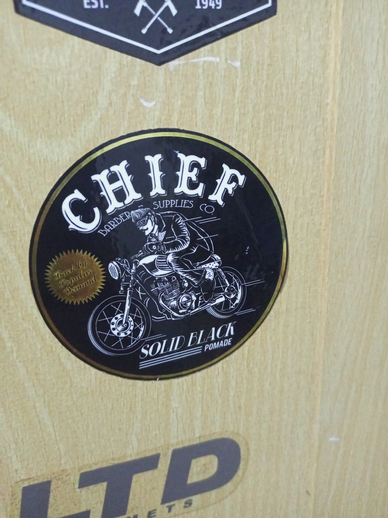 A circular black sticker with a gold border is affixed to a light-colored wooden surface. The sticker features the word "CHIEF" in large, stylized white lettering at the top, followed by "BARBER SUPPLIES CO." in smaller white lettering. In the center of the sticker is a black and white illustration of a man on a motorcycle. The man is wearing a jacket, sunglasses, and has a slicked-back hairstyle. To the left of the illustration, a gold sunburst shape contains the text "Back by Popular Demand" in cursive. At the bottom of the sticker, the words "SOLID BLACK" are in white capital letters, with "POMADE" below it in smaller white lettering. A partial view of another sticker with large gray letters is visible at the bottom of the image. The wooden surface appears to be a wall or a piece of furniture.