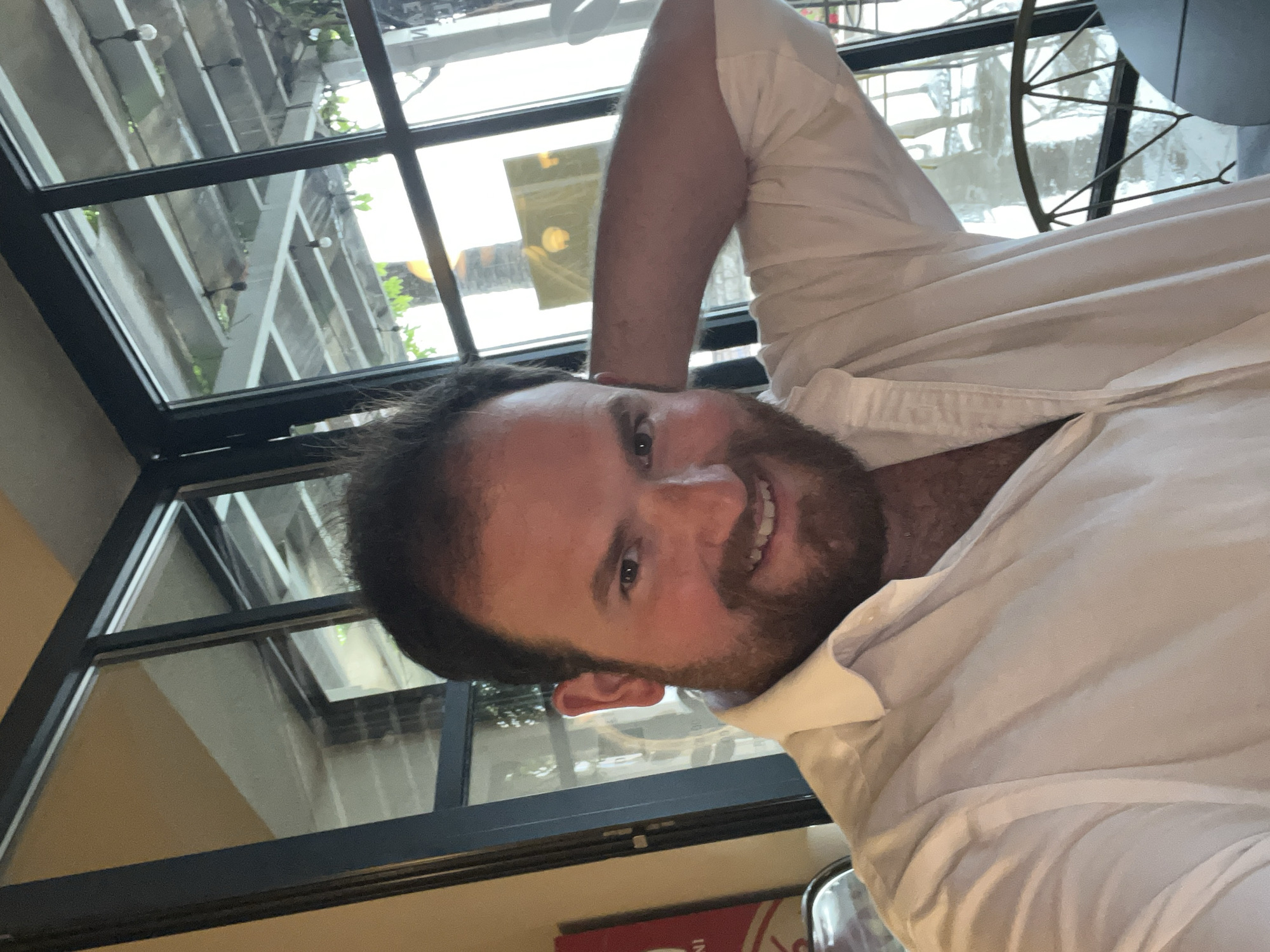 A man with dark hair and a beard smiles towards the camera. He is wearing a white shirt, and his arm is raised and bent behind his head. Behind him is a window with multiple panes, showing an outdoor scene with a partially visible pergola or similar structure. The outdoor scene appears to be in Rafailovići, Montenegro, based on the architectural style. A corner of a red and white object is visible in the lower part of the frame, possibly a menu or placemat. The lighting suggests an indoor setting, potentially a restaurant or cafe.