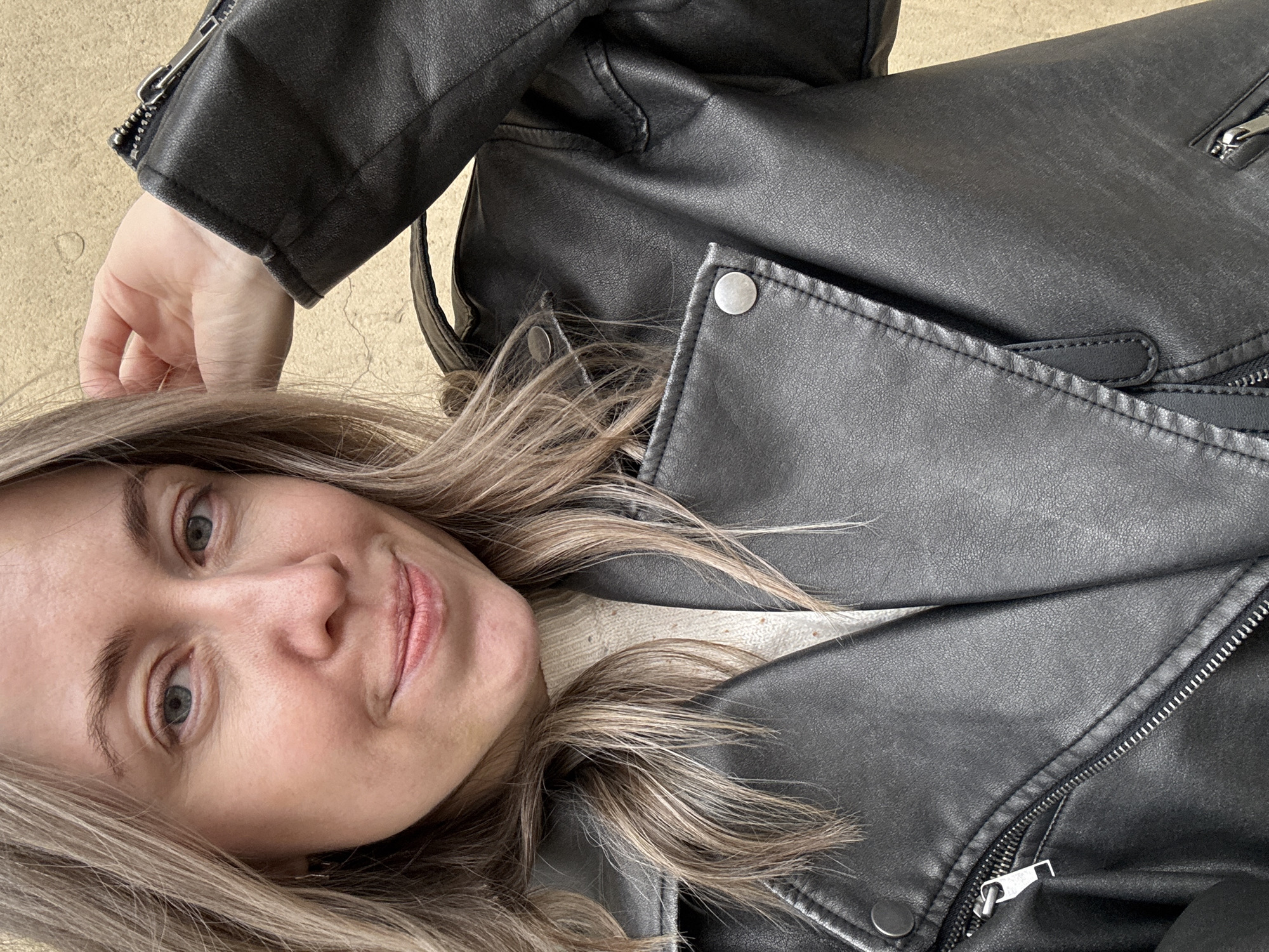 A woman with blonde and light brown hair poses close to the camera, smiling slightly with light pink lips. She has fair skin and light-colored eyes. She is wearing a black leather jacket, which dominates the upper portion of the frame. The jacket has visible zippers, snaps, and stitching details. The background appears to be a light beige or tan surface. The photograph does not reveal any elements that would indicate the location is Saint Petersburg, Russia.