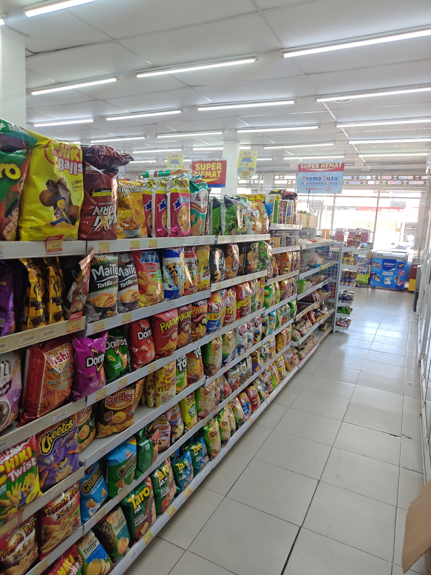 The image shows an aisle in a convenience store in Klampokan, Indonesia, filled with shelves of various snack products. The shelves are stocked with numerous bags of chips, crackers, and other packaged snacks, predominantly with colorful packaging. Prominent brands like Cheetos, Doritos, and Taro are visible. The store appears well-lit with fluorescent lighting fixtures on the ceiling. Signs indicating "SUPER HEMAT" (Super Savings) and "Promo Susu" (Milk Promo) are displayed, suggesting a promotional event. In the background, a refrigerator unit with ice cream products and other packaged goods can be seen. The floor is tiled, and a cardboard box is partially visible in the lower right corner. There are no people visible in the image.