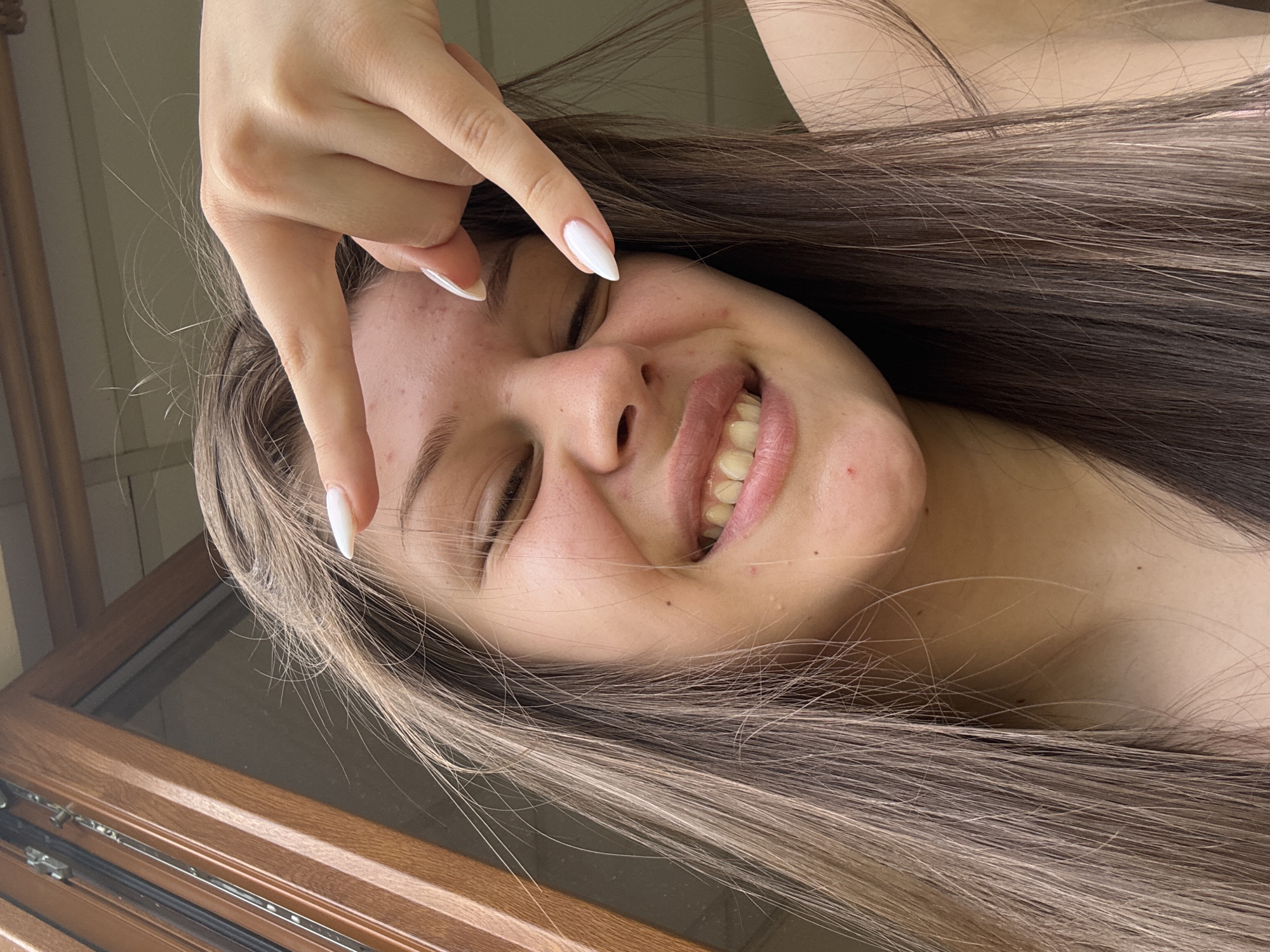 A young woman with long, dark brown hair is looking at the camera and smiling with her eyes closed. Her right hand is raised, with her index finger pointing towards her forehead. She has white nail polish on her fingernails. The background appears to be indoors, with a wooden window frame on the left side of the image. The lighting suggests it might be daytime. The woman has some acne on her forehead and cheeks.