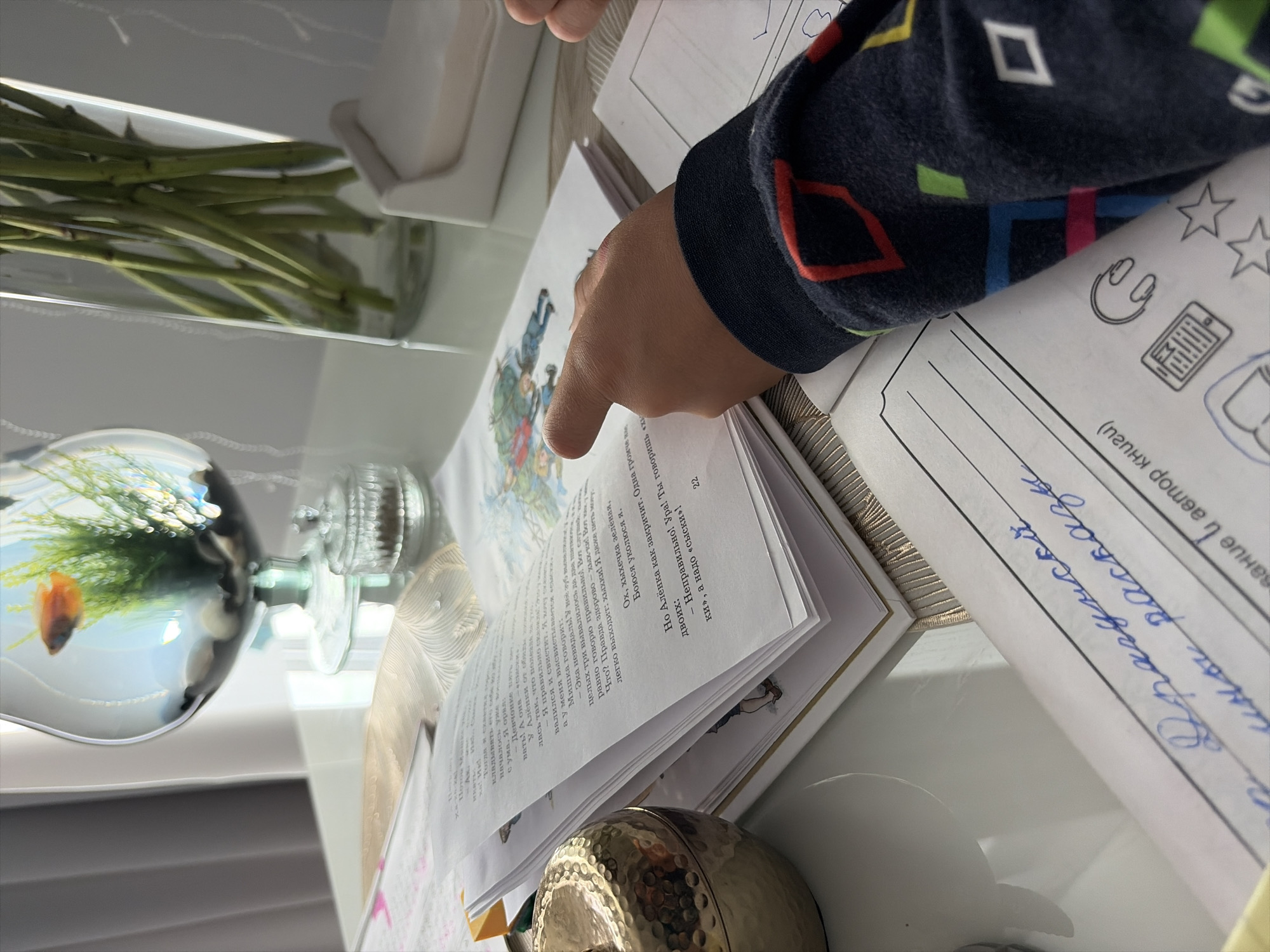 A close-up, indoor shot depicts a table with an open book and a child's hand pointing at a page. The book appears to be a children's storybook with illustrations. To the left, a decorative, golden, apple-shaped object sits on the table. In the background, a fishbowl with a single fish and aquatic plants is visible, along with a tall glass vase containing green stems, possibly flowers, and a small, clear glass container. The scene is set on a reflective white surface, likely a table, which is draped with a textured, light-colored fabric. The lighting suggests a well-lit indoor environment, possibly a home. The text within the book and on other papers on the table is in Cyrillic script. Based on the Cyrillic script, this scene is likely located in Moscow, Russia.