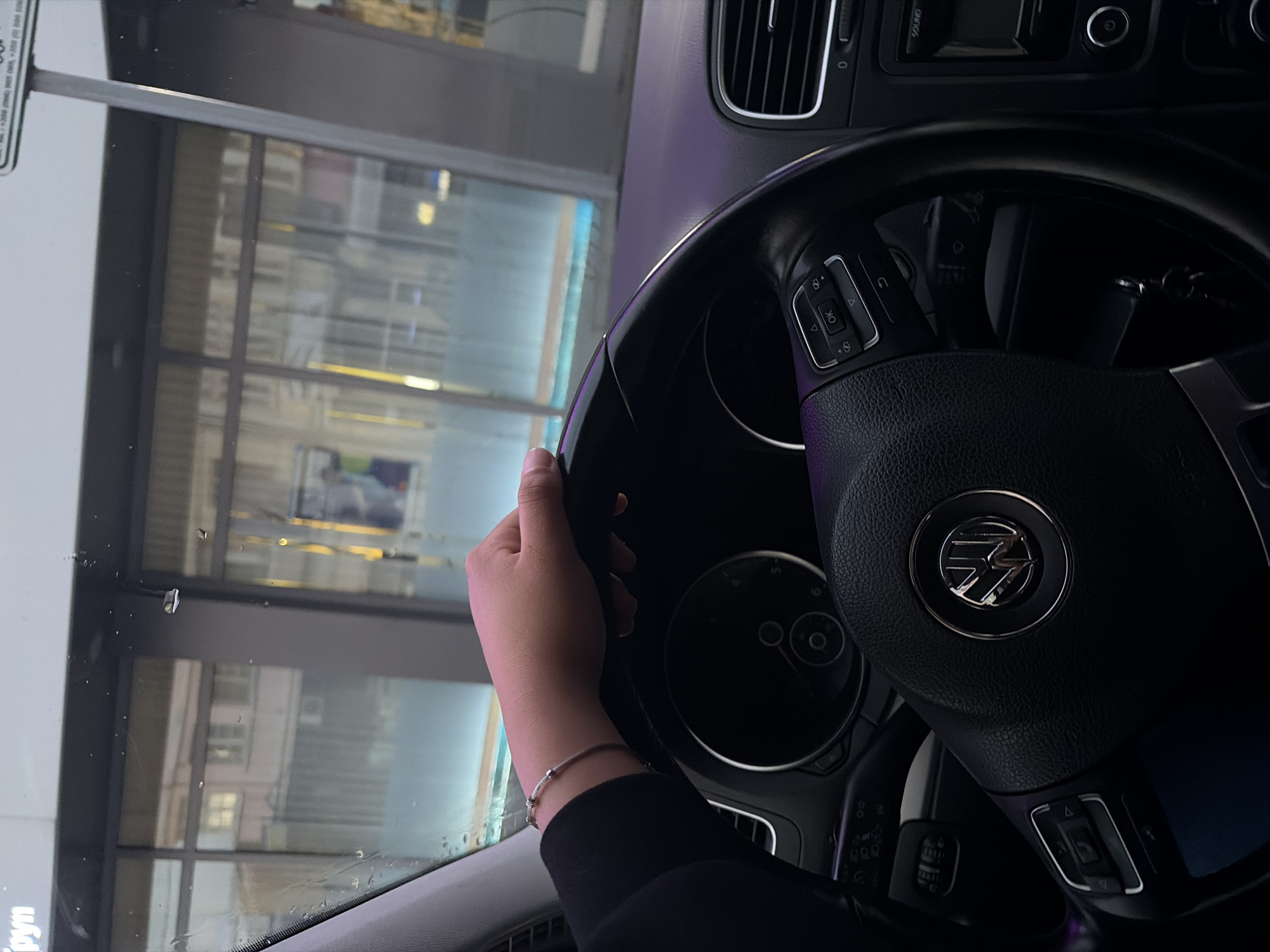 The image shows the interior of a car from the driver's perspective, with a focus on the steering wheel and the view through the windshield. A human hand, adorned with a silver-colored bracelet, is holding the upper-left portion of the black, leather-wrapped steering wheel. The steering wheel prominently displays the Volkswagen logo in its center, along with various control buttons including "OK" and directional arrows.
Part of the car's dashboard is visible, featuring an air vent, and controls for the infotainment system, with "SOUND" visible above one knob. The scene outside the car, viewed through the windshield, appears to be an urban or commercial environment during the evening or night, indicated by prominent reflections of artificial lights and architectural elements with large windows.
A white sign or board is visible to the left through the windshield, with some indistinct text and numbers partially reflected, including fragments like "...PM" and "...Tagen". The windshield has some smudges or possibly small water droplets. The overall setting suggests the car is either parked or stopped, and the location context provided is Gabrovo, Bulgaria.