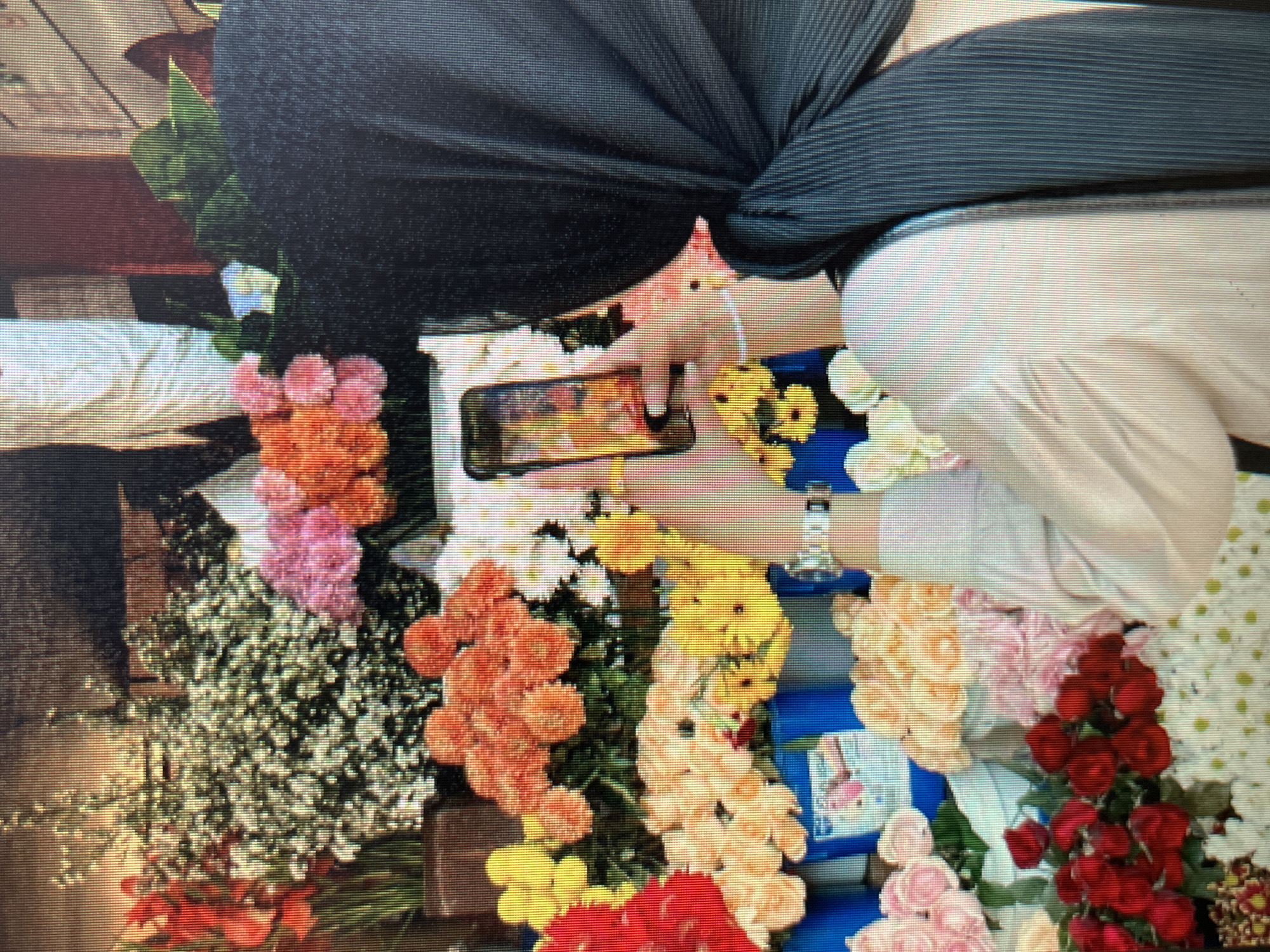 The image depicts a street-level view of a flower stall in Sejahtera, Indonesia. A person wearing a black hijab and a light pink shirt is taking a photograph of the flowers with their smartphone. The stall is filled with a vibrant assortment of flowers, including red gladiolus, an abundance of small pink and orange carnations, yellow daisies, and bunches of pale yellow and peach roses. Blue buckets are visible holding some of the flower arrangements. The background is slightly blurred, but suggests a busy urban environment.