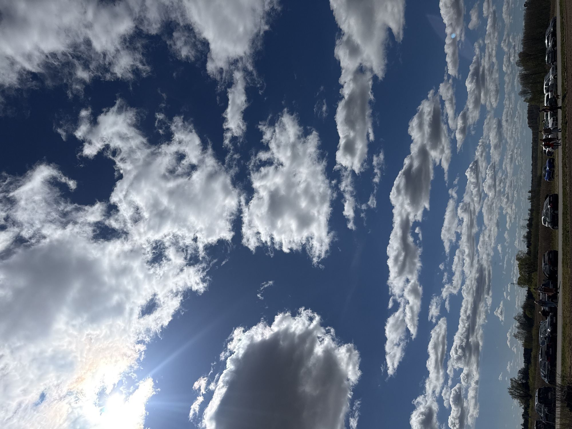 The image depicts a bright, partly cloudy sky with a deep blue background. The sun is visible, with its rays shining through the clouds. On the right edge of the image, there is a row of parked cars next to a line of trees, suggesting a location near the side of a road or a parking area. Given the context of Beitiņi, Latvia, it is plausible this is a rural roadside stop, or the edge of a parking lot at a park or rural venue near Beitiņi. There is no way to confirm without more information. There are also possible people at the roadside but they are too far away to be discernible.