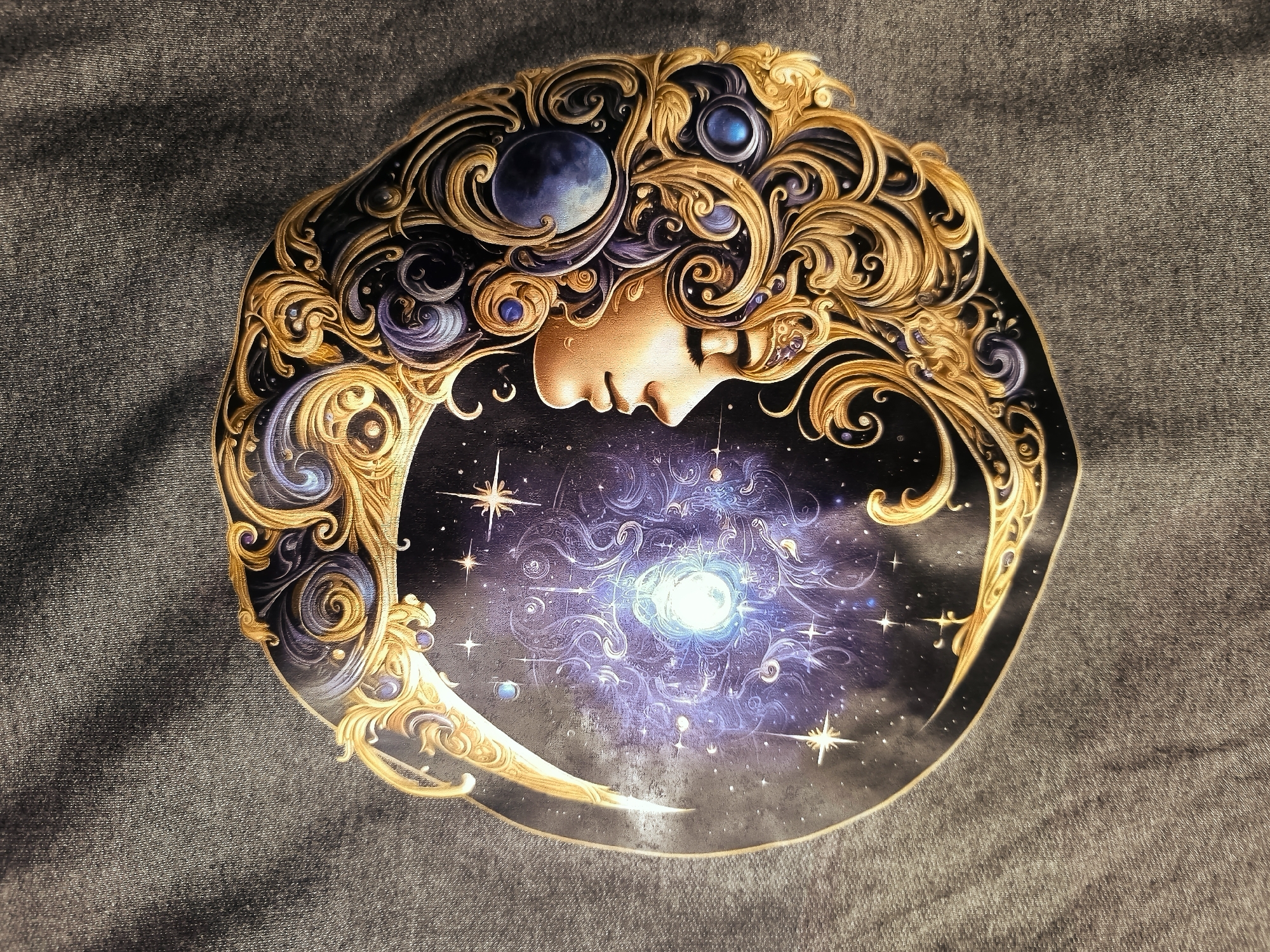 The image displays a circular artwork with a celestial theme, resting on a dark gray, textured fabric surface, possibly denim. The artwork depicts a profile of a woman's face with closed eyes, her hair interwoven with ornate gold and dark blue swirling patterns that resemble nebulae and cosmic dust. Embedded within her hair are several celestial bodies, including a blue moon and small, glowing blue gems. The background of the artwork is a starry night sky, featuring a bright, glowing central light source with swirling cosmic patterns around it, suggesting a vibrant nebula or star cluster. The outer edge of the artwork is framed by elaborate gold scrollwork.
The location in the city of Almere, Netherlands, is not discernible from the provided image. There are no visible landmarks, street signs, or environmental clues that can identify a specific location within Almere.