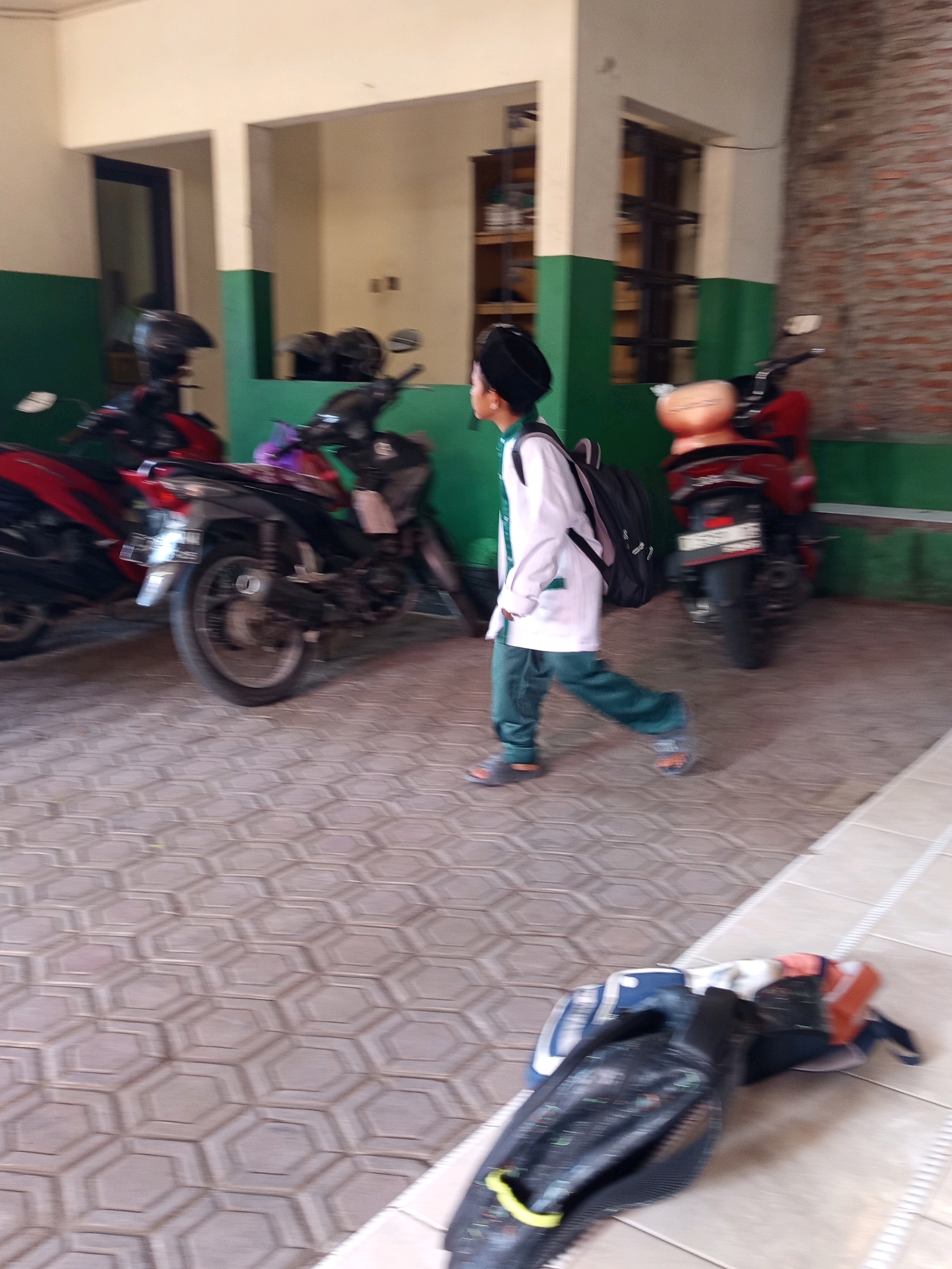 The image depicts an outdoor scene, likely on school grounds in Karawang, Indonesia, with several motorcycles parked. A young person, dressed in a white shirt, green pants, and a black head covering, is walking from left to right across a tiled courtyard. They are carrying a black backpack. In the background, there are more motorcycles and a brick wall. Two bags are visible on the ground in the foreground.