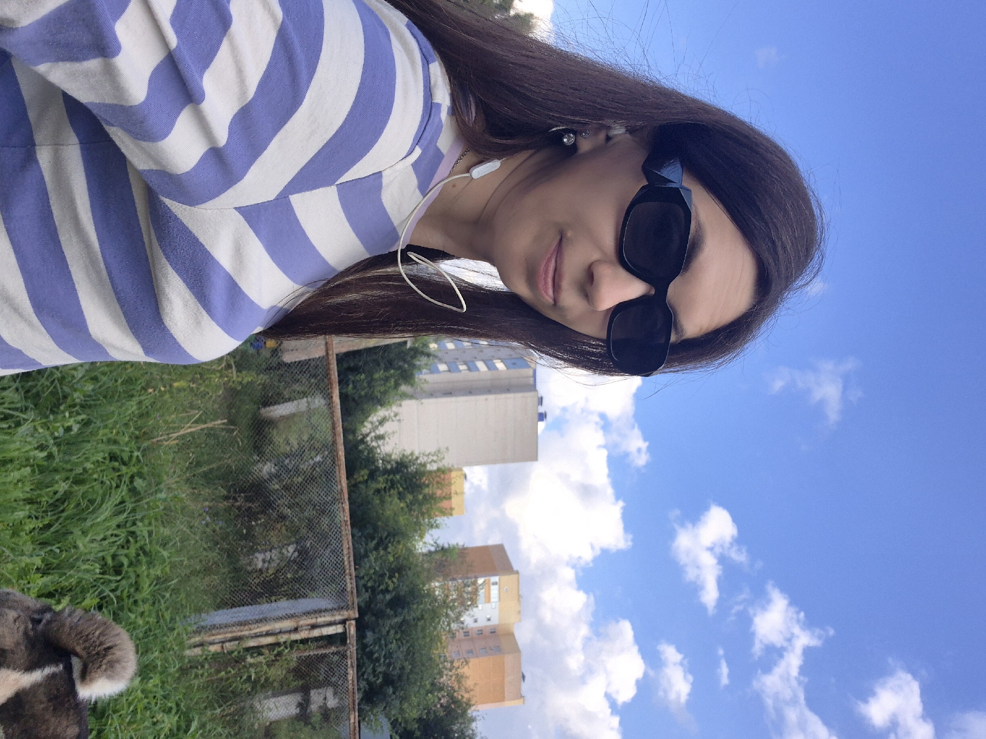 A woman is taking a selfie outdoors. She has long dark hair and is wearing large black sunglasses and a purple and white striped shirt. She has white earbuds in her ears and a white cord from the earbuds is visible around her neck. The background shows a blue sky with white clouds, tall apartment buildings, and trees. A chain-link fence is also visible in the middle ground, and some tall green grass and a dog are in the foreground. The woman is smiling slightly. The scene is consistent with a residential area in Chaikovsky, Russia, given the architectural style of the buildings and the presence of greenery.