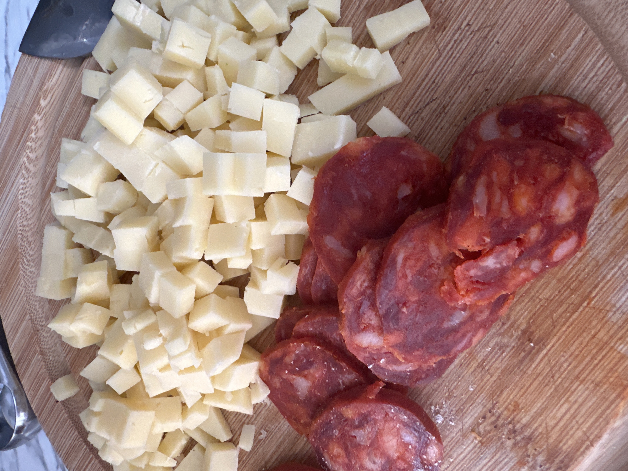 The image shows a close-up, overhead shot of diced cheese and sliced chorizo on a wooden cutting board. The cheese is light yellow and cut into small, irregular cubes, piled in the upper right portion of the frame. The chorizo slices are circular with a deep red color and visible white fat marbling, arranged in the lower left of the image. The wooden cutting board has a visible grain pattern and some scattered crumbs. In the upper left corner, a small portion of a metal utensil is visible. There are no people or indications of a city location.