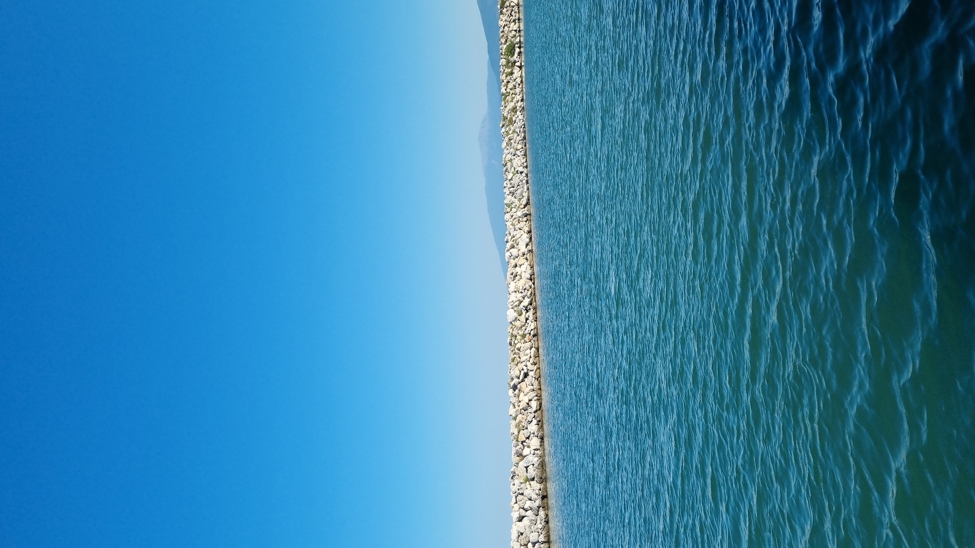 The image shows a wide expanse of calm, blue-green water in the foreground. A breakwater made of large, light-colored stones stretches horizontally across the middle ground, separating the water from a distant, hazy shoreline. The sky above is a clear, bright blue, with no clouds visible. In the far distance, a faint outline of mountains can be seen against the horizon. The scene appears to be a natural or semi-natural coastal area, possibly a harbor or a protected bay. The location is identified as Limani Litochorou, Greece. There are no people or visible signs of activity in the image. The overall impression is one of tranquility and open space.