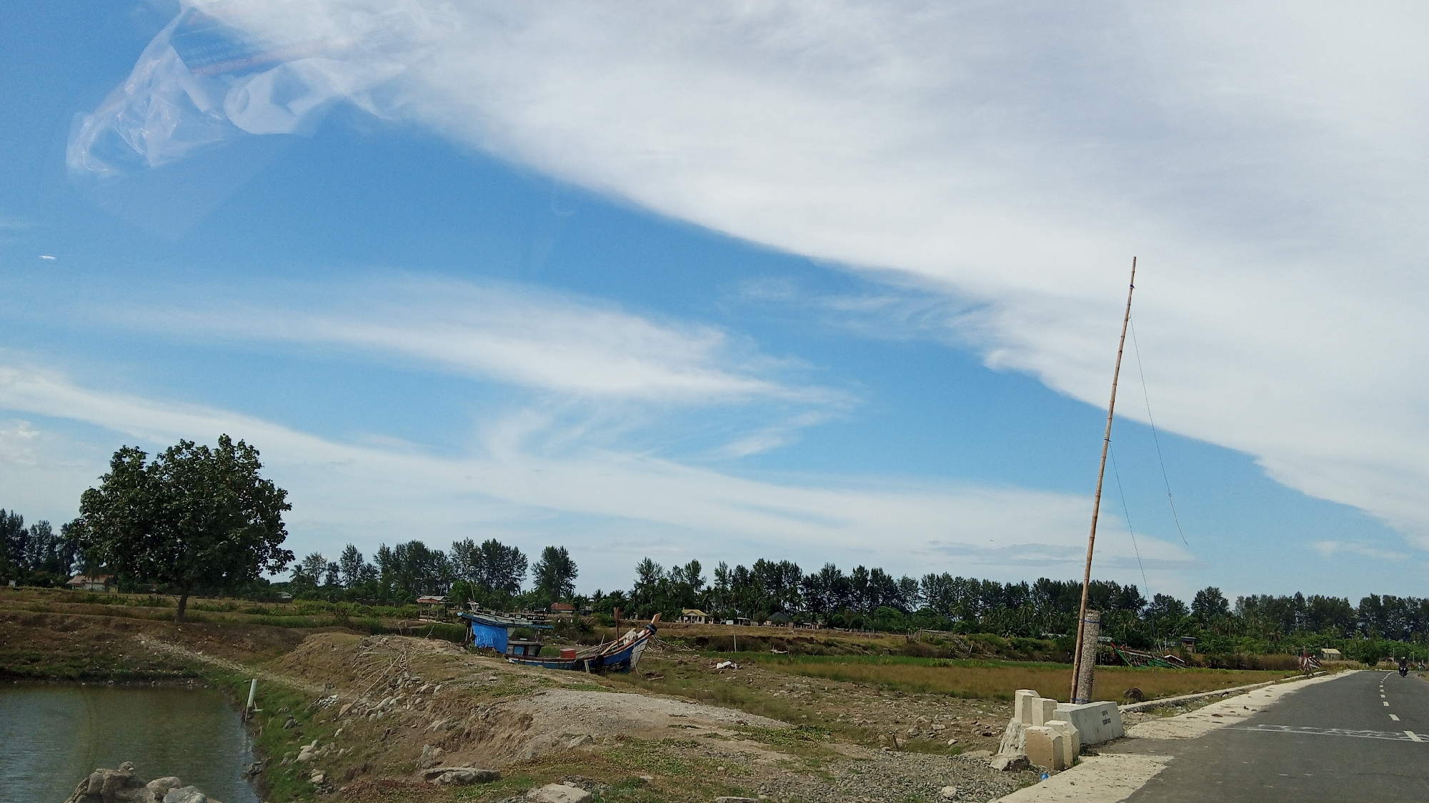 The image depicts a rural landscape with a dirt road on the right side, curving into the distance. A single motorcyclist is visible on the road. In the foreground, there is a body of water and a rocky embankment. A large tree stands to the left of the frame, with a line of trees forming a dense background. Several small boats, some with blue tarps, are moored near the water's edge. A tall bamboo pole with wires attached stands vertically in the middle ground. The sky is bright blue with scattered white clouds. The scene appears to be located in a coastal or riverside area, characteristic of Tanjong Krueng, Indonesia.