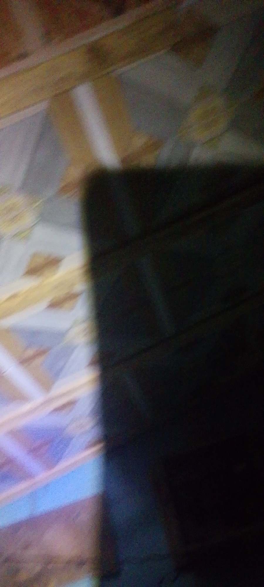 This media file is an abstract, low-resolution image that primarily features a dark, indistinct object occupying the right half of the frame. The left side of the image displays a blurry, patterned surface that appears to be flooring or a tiled wall. The pattern consists of diagonal lines and geometric shapes in muted tones of beige, yellow, and light blue, suggesting a decorative element.
The overall impression is one of dim lighting and a lack of clear focus. The dark object on the right is a solid mass with subtle variations in shading, hinting at a textured surface or folds, but its identity is not discernible. There are no visible people, text, or specific environmental cues to determine the time of day, weather, or exact location within Jalingo, Nigeria. The image lacks any discernible activity or narrative, existing as a collection of abstract forms and colors.