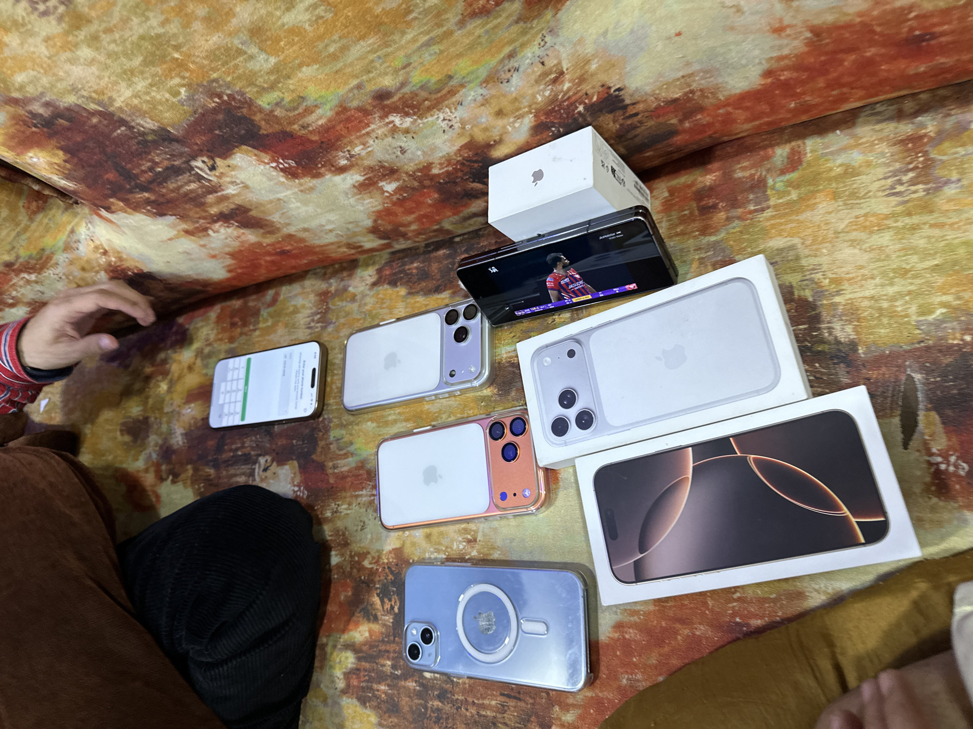 The image shows several iPhones, some with cases, laid out on a patterned surface. One iPhone displays a numeric keypad on its screen. Another iPhone sits atop an Apple box and shows a soccer game on its screen. The scene appears indoors on an area that could be a sofa, with part of a person visible on the left. The location in Shahapur, India, is unspecified within the visual details of the photograph.