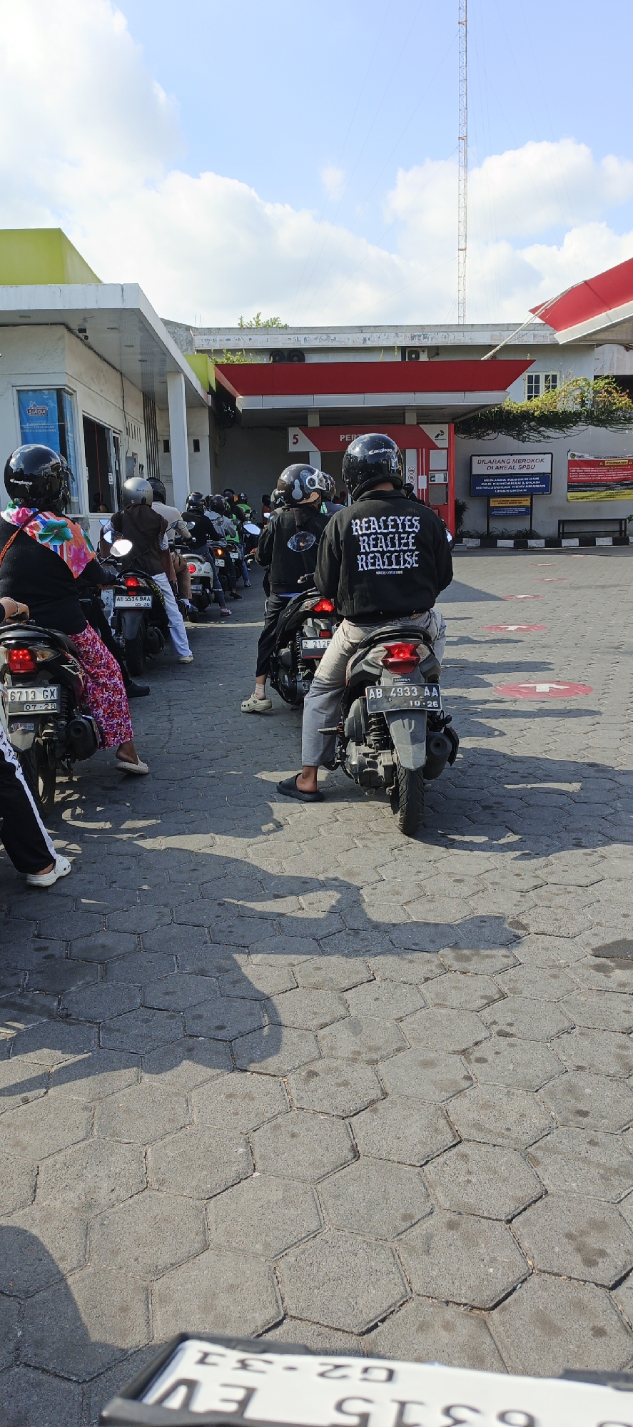 The image captures a queue of motorcyclists at a gas station under a bright, partially cloudy sky. The gas station features a red and white awning extending over multiple pump stations. Signs with Indonesian text, including "DILARANG MEROKOK DI AREA SPBU" (No smoking in the gas station area), are visible on a white wall. A tall communication tower stands in the background. The ground is paved with hexagonal concrete tiles. Several motorcyclists are wearing helmets, and one in the foreground has a black jacket with white text that reads "REALEYES REALIZE REALISE." License plates visible on motorcycles include "AB 5534 BA," "6713 GX," and "AB 4933 AA." The overall scene suggests a typical day at a busy gas station in Kasihan, Indonesia, with people waiting to refuel their vehicles.