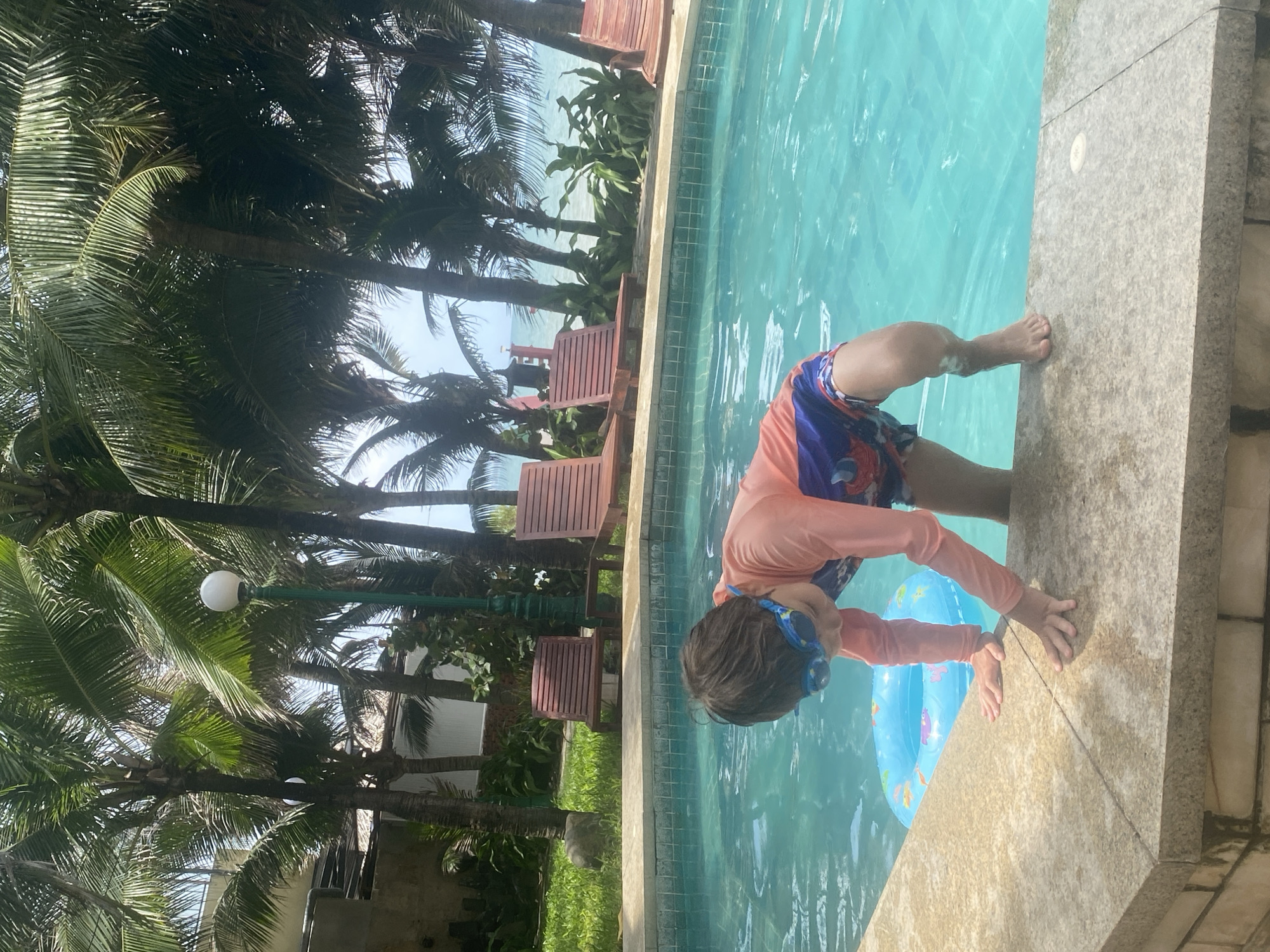 A young child wearing blue swim goggles and a long-sleeved pink rash guard over patterned swim trunks is positioned at the edge of a turquoise swimming pool, with their hands braced on the pool coping and one foot submerged in the water. A blue inflatable ring with colorful fish graphics floats in the pool near the child. The background features palm trees, lounge chairs, and a glimpse of the ocean under a bright sky. This scene is located in Phu Thuy Ward, Vietnam.