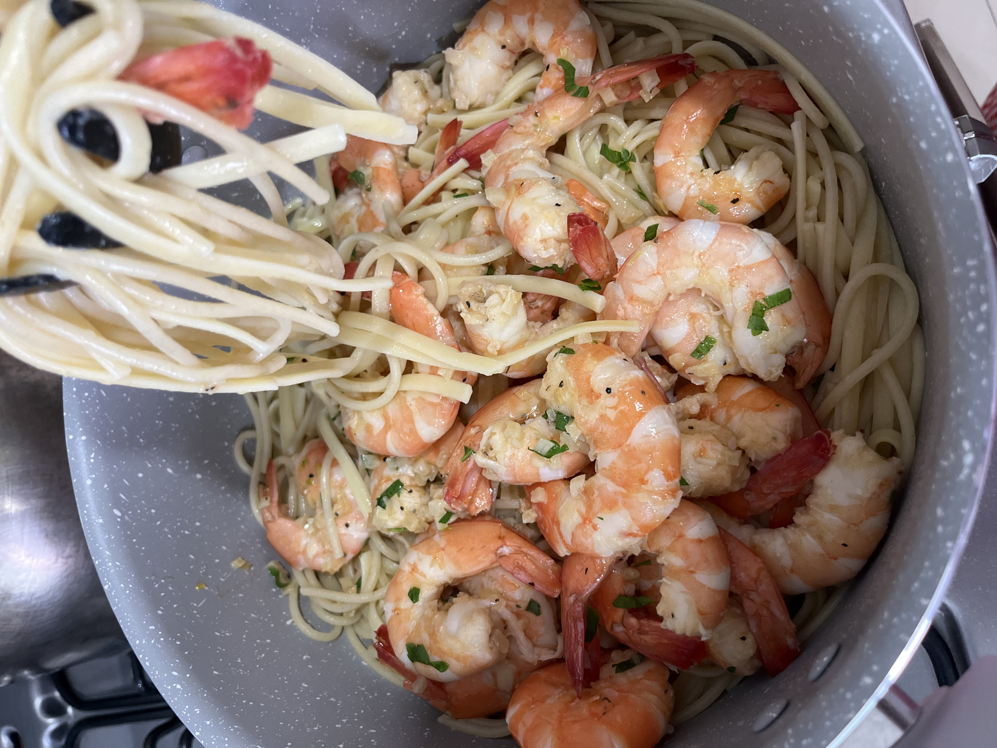 The image depicts a close-up view of a pot filled with linguine pasta and shrimp scampi. A serving utensil with linguine, olives, and tomatoes is held above the pot. The shrimp are scattered throughout the pasta, and are of medium size and appear to be seasoned with herbs. The pot is gray and white. The location is Barrio de Nuevo León, Mexico.