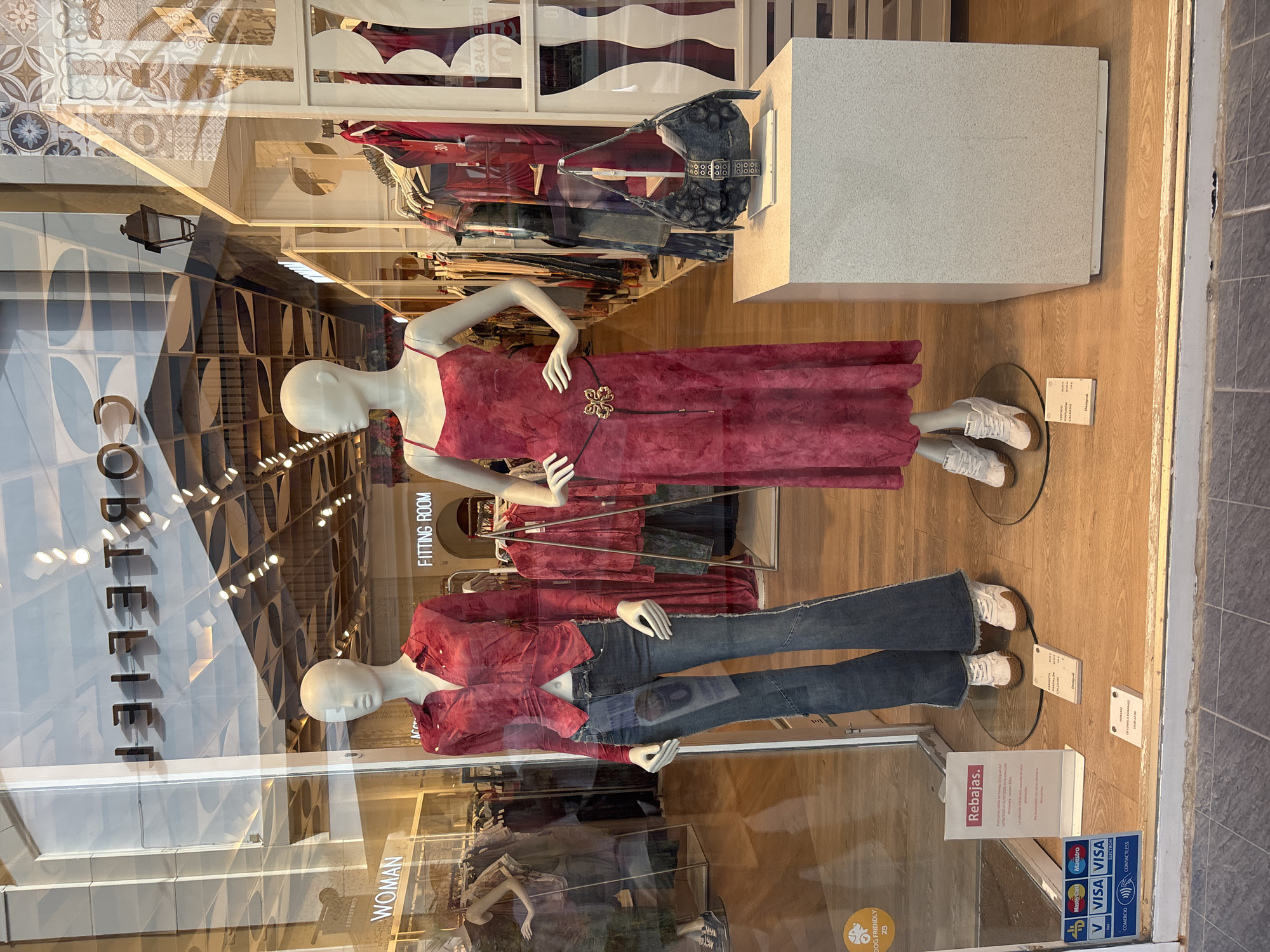 This is a full-shot, eye-level exterior photograph of a clothing store window display on a sunny day. The storefront is a glass window revealing the interior of the store. The window is clean and clear, reflecting the exterior surroundings, including trees and the sky.
Inside the window, two mannequins are displayed. The mannequin on the left is wearing a red tie-dye blouse and blue flared jeans. The mannequin on the right is wearing a long, pink tie-dye dress with a black belt that has a gold butterfly buckle. Both mannequins are wearing white sneakers with brown soles.
In front of the mannequin on the right, a gray patterned handbag is displayed on a light-colored pedestal. Behind the mannequins, shelves of clothing are visible, along with a sign that says "WOMAN" and another that says "FITTING ROOM".
A sign on the lower left side of the window displays payment icons (Visa, Mastercard, Maestro, VPay, Electron) and a "DOG FRIENDLY" sticker. Another sign in the lower center of the window reads "Rebajas" (Sales) and provides details of a promotion. Additional signage in the bottom right corner indicates store hours. The flooring inside the store appears to be wood. The storefront is located in the city of Orihuela, Spain.