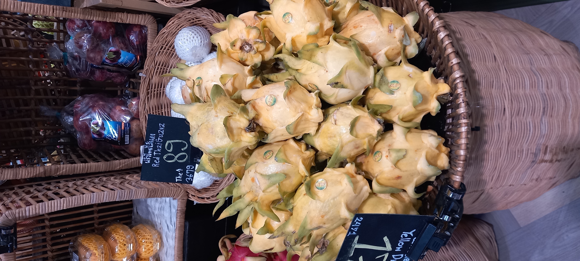 The image captures a vibrant display of fresh produce arranged in woven wicker baskets, likely within a market or grocery store in Moo 1, Thailand. The scene is dominated by a large quantity of yellow dragon fruit (pitaya) piled in a central basket. These fruits are characterized by their bright yellow skin and green-tipped scales, with several bearing small green stickers.
To the left, on a lower shelf within a wicker structure, are clear plastic bags containing passion fruit. Labels on these bags clearly show the text "FRESH Passion" with "fruit" partially visible. Nearby, round fruits wrapped in white mesh netting are also visible, possibly red Thai guavas.
Two black price tags are prominent in the foreground. One tag, positioned near the yellow dragon fruit, displays "Yellow D" (likely for Yellow Dragon Fruit) and the number "2471". The other tag, to the left, features Thai script "ฝรั่งไทยไส้แดง" along with the English translation "Red Thai Guava". Below this, "The 89" suggests a price of 89 (presumably Baht), followed by the number "3679".
The arrangement suggests a retail setting where fruits are presented for sale. The use of natural wicker baskets adds to the aesthetic appeal of the fresh produce display. The lighting is bright and even, highlighting the colors and textures of the fruits. No people are visible, and the time of day or weather cannot be determined from the image.