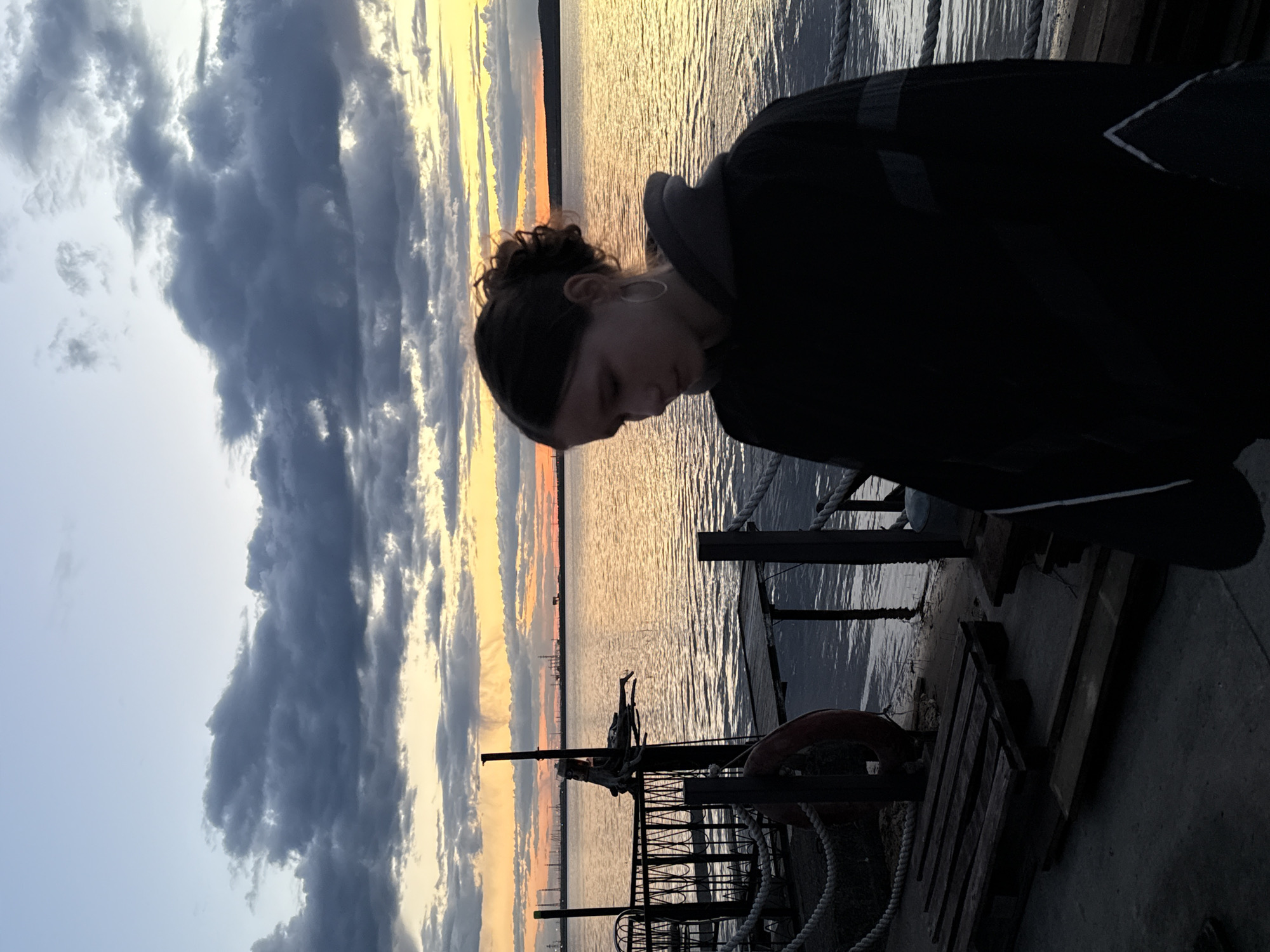 A low-angle, outdoor shot captures a person standing on a pier overlooking a body of water. The person, likely female, has their head bowed and is wearing a dark hoodie with a hood. A hair band can be seen. The pier is made of wood and concrete, with a metal railing and a life preserver visible. The background displays a dramatic sunset with dark clouds and golden light reflected on the water. The location is likely in the city of Chaikovsky, Russia, given the prompt. There is a city skyline visible in the far distance of the sunset.
