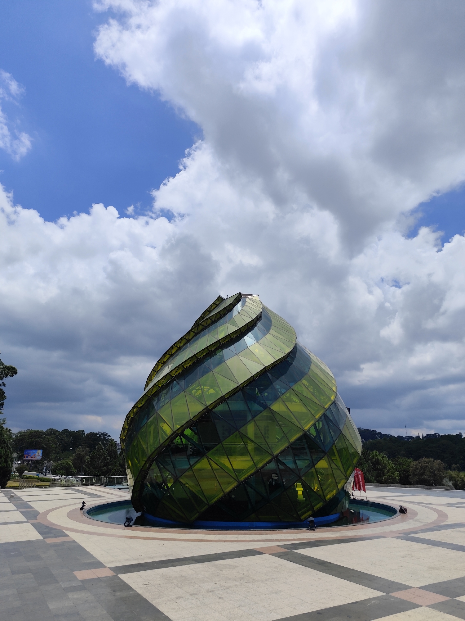 The image shows a large, spiraling, glass-paneled structure resembling an artichoke. This structure is located in a plaza in the city of Da Lat, Vietnam, surrounded by a shallow pool of water. The plaza has a patterned, tiled floor. In the background, there is vegetation and greenery, suggesting a park or landscaped area. The sky is partly cloudy, with patches of blue sky visible. There are a few small figures (possibly people) visible near the base of the glass structure, but they are not clearly identifiable. There are also two red flags next to the building.