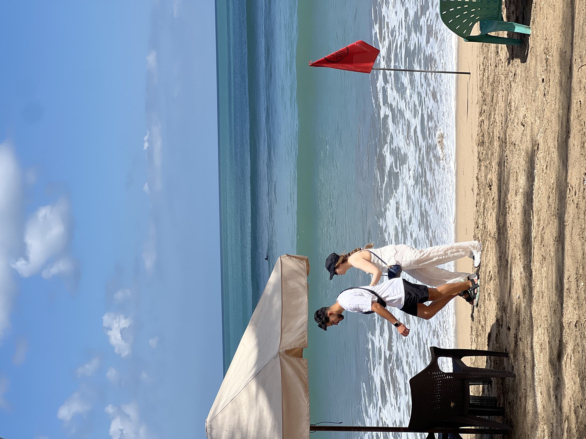 The image depicts a beach scene in Kuta, Indonesia, under a bright, partly cloudy blue sky. In the foreground, a beige beach umbrella shades two plastic chairs, one of which is dark brown and the other is green. A young couple is walking along the sandy shore. The man, wearing a white t-shirt and black shorts, has a black fanny pack across his chest. The woman, in a white dress and a black cap, carries a small blue bag. The ocean in the background displays waves of varying shades of blue and turquoise, with white foam breaking on the shore. A red flag on a pole stands near the water's edge. A lone surfer is visible in the distance on the water. The sand on the beach is light brown, with some patches of shadow cast by the umbrella. The scene conveys a relaxed, sunny day at the beach.