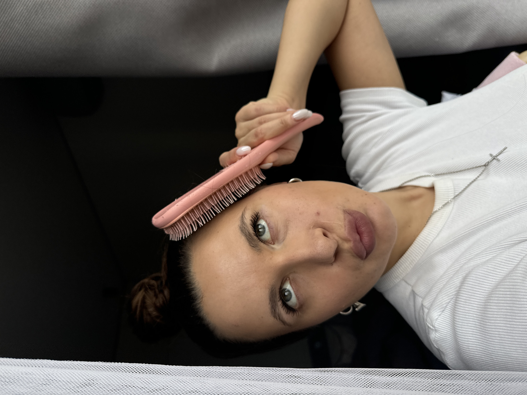A woman is lying down, holding a pink hairbrush set vertically above her forehead. She is wearing a white shirt and a silver necklace with a cross pendant. She has light-colored eyes, dark hair pulled back in a bun, and white nail polish. The background appears to be a combination of gray and black surfaces. The location in the city of Akulovo (Gp Kubinka), Russia, is not discernible from the visual information provided.