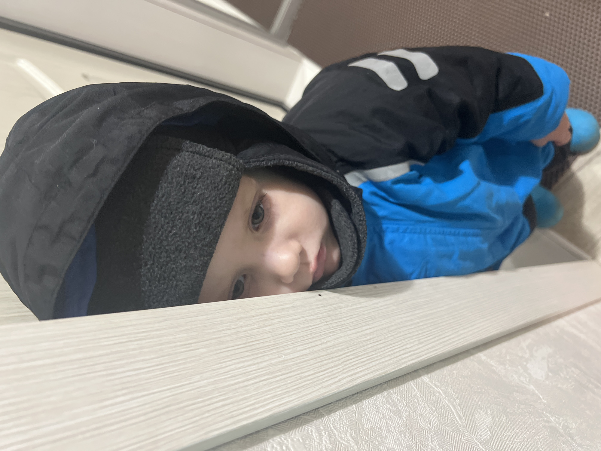 A young child wearing a black and blue hooded jacket lies on a white surface that appears to be a counter or table. The child's face is visible, and they are looking towards the camera. The background is out of focus but shows a room interior. No information confirms the location being Марковский, Russia.
